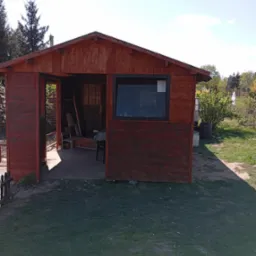 Drewniana altana ogrodowa z oknem i otwartymi drzwiami, pomalowana na brązowo, otoczona zielenią. Widoczne elementy wyposażenia wewnątrz.