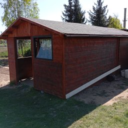 Mirosław Gielka - Ciemnobrązowa drewniana altana z oknem i otwartą przestrzenią, widoczna na trawniku. Elementy konstrukcyjne w trakcie montażu. W tle drzewa i błękitne niebo.