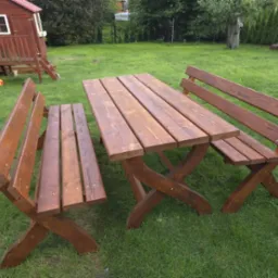 Drewniany stół piknikowy z dwiema ławkami na trawniku, w tle domek dla dzieci i drzewa. Widoczne detale konstrukcyjne i faktura drewna.