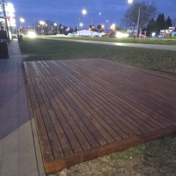 Mirosław Gielka - Nowoczesny taras z ciemnego drewna, ułożony na poziomie gruntu, z widocznymi śladami użytkowania. Tło stanowi trawnik i oświetlona ulica w scenerii zmierzchu.
