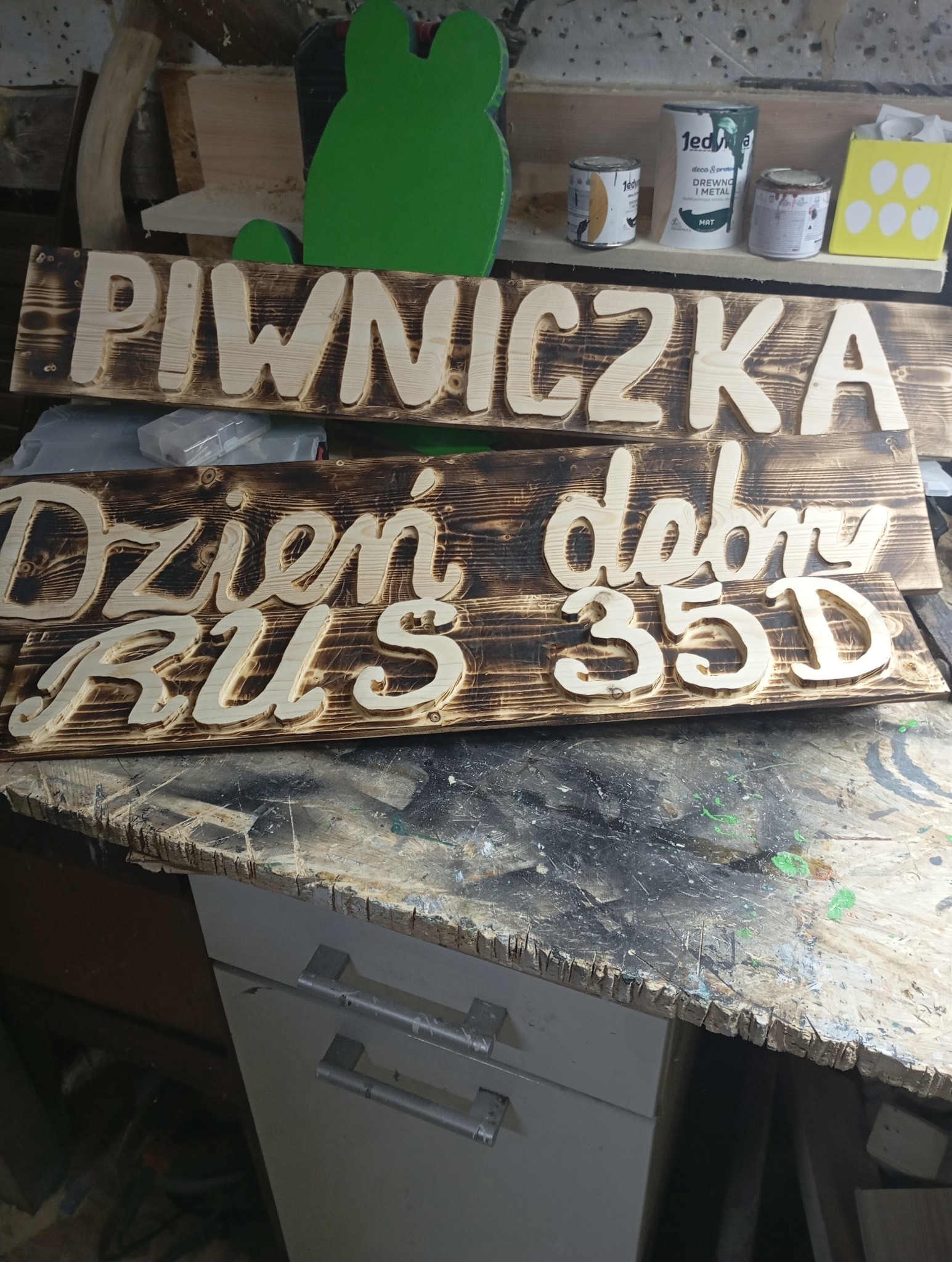 Dwie drewniane tabliczki z wyciętymi napisami 'Piwniczka' i 'Dzień dobry RUS 35D', z wypalonym tłem, leżące na warsztatowym stole, w tle puszki z farbami i zielona wycinanka w kształcie żaby.