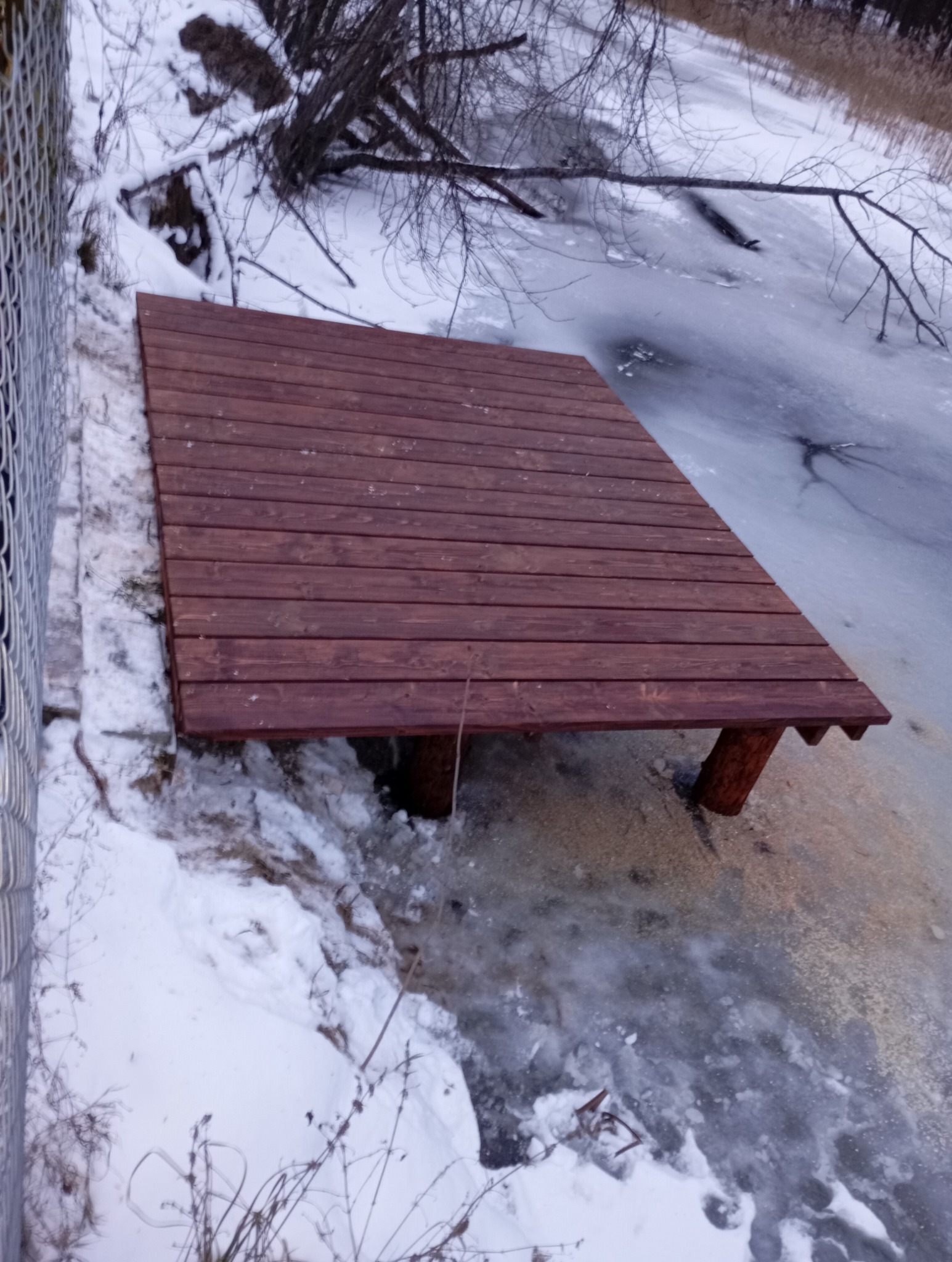 Drewniany taras w Świątkach, zbudowany nad częściowo zamarzniętym brzegiem rzeki, otoczony śniegiem i siatką ogrodzeniową.