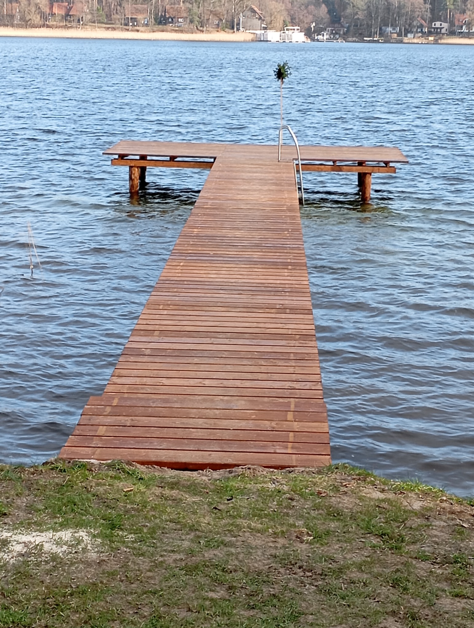 Długi, drewniany pomost wchodzący w jezioro, zakończony platformą z metalową drabinką, w tle zalesiony brzeg.