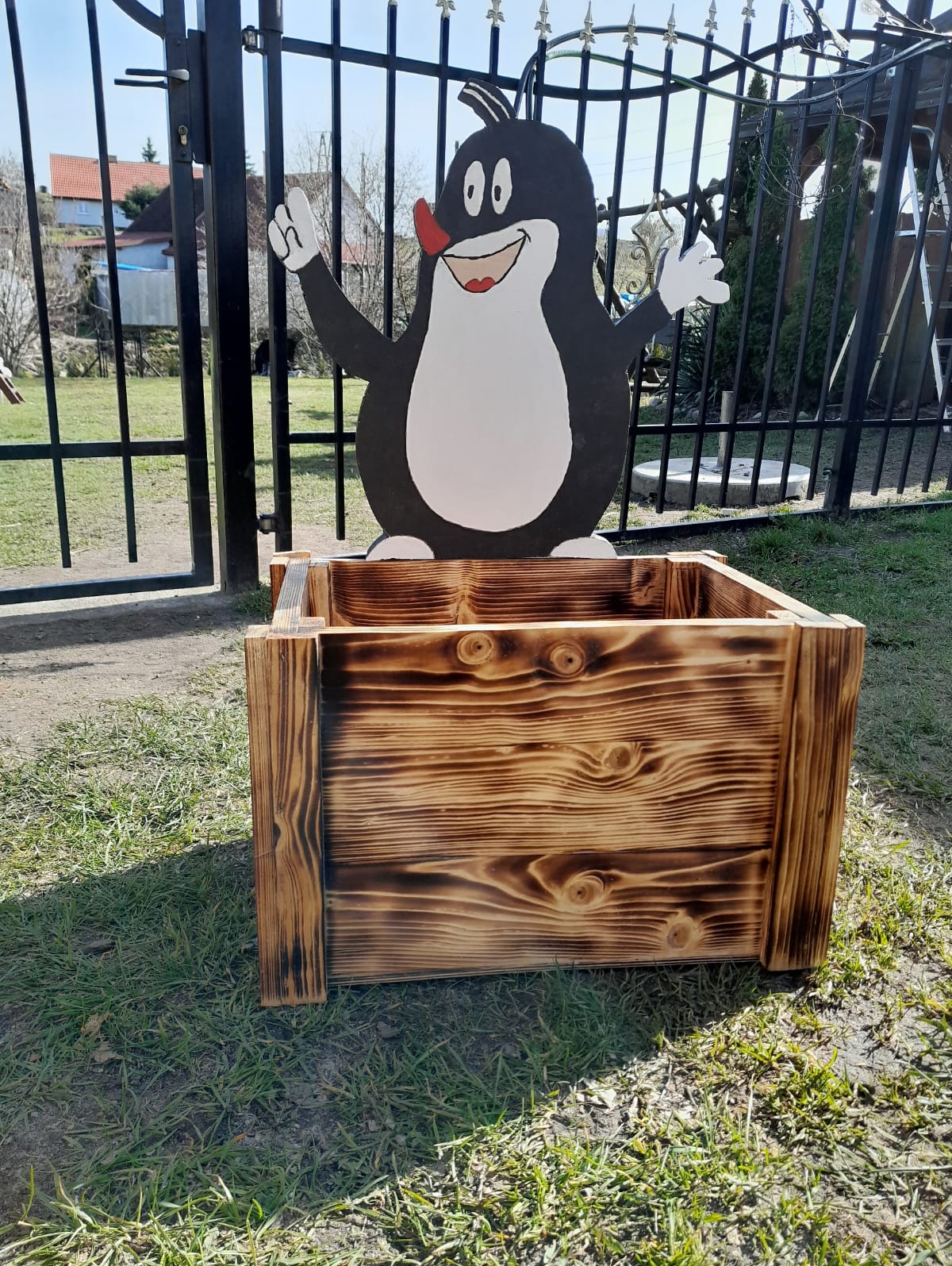 Opalana drewniana skrzynia na rośliny z dekoracją w postaci postaci Krecika, umieszczona na trawniku przed metalowym ogrodzeniem.
