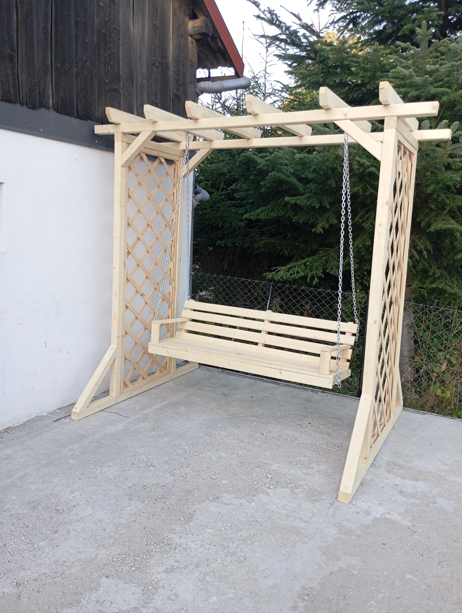 Drewniana huśtawka ogrodowa z kratownicowymi bokami, zawieszona na łańcuchach, ustawiona na betonowym podłożu przed ścianą domu i zielonymi drzewami.