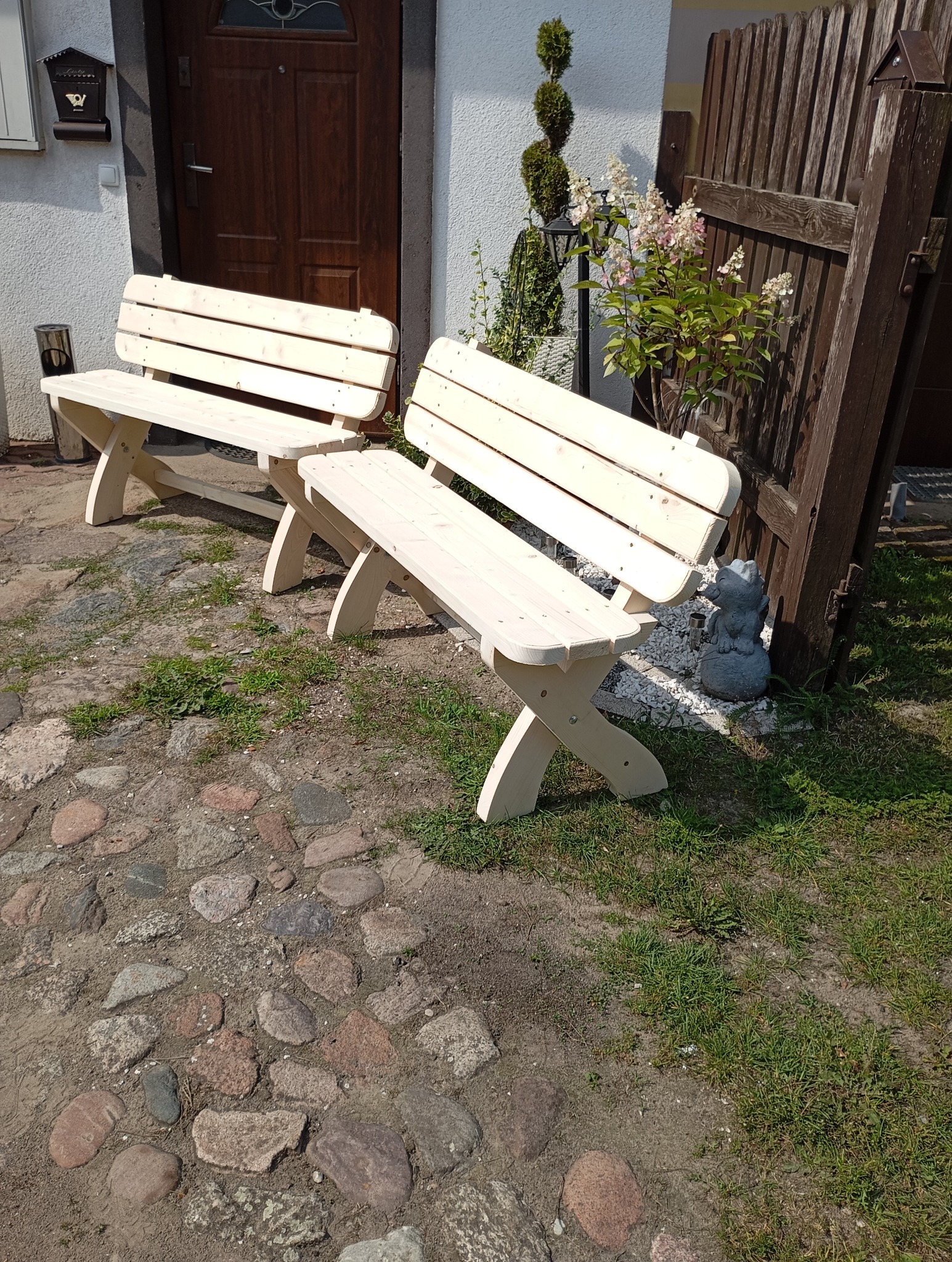 Dwie surowe, jasne, drewniane ławki ogrodowe z krzyżowymi nogami stoją na brukowanym podwórku przed domem z ciemnymi drzwiami i ozdobnymi roślinami.