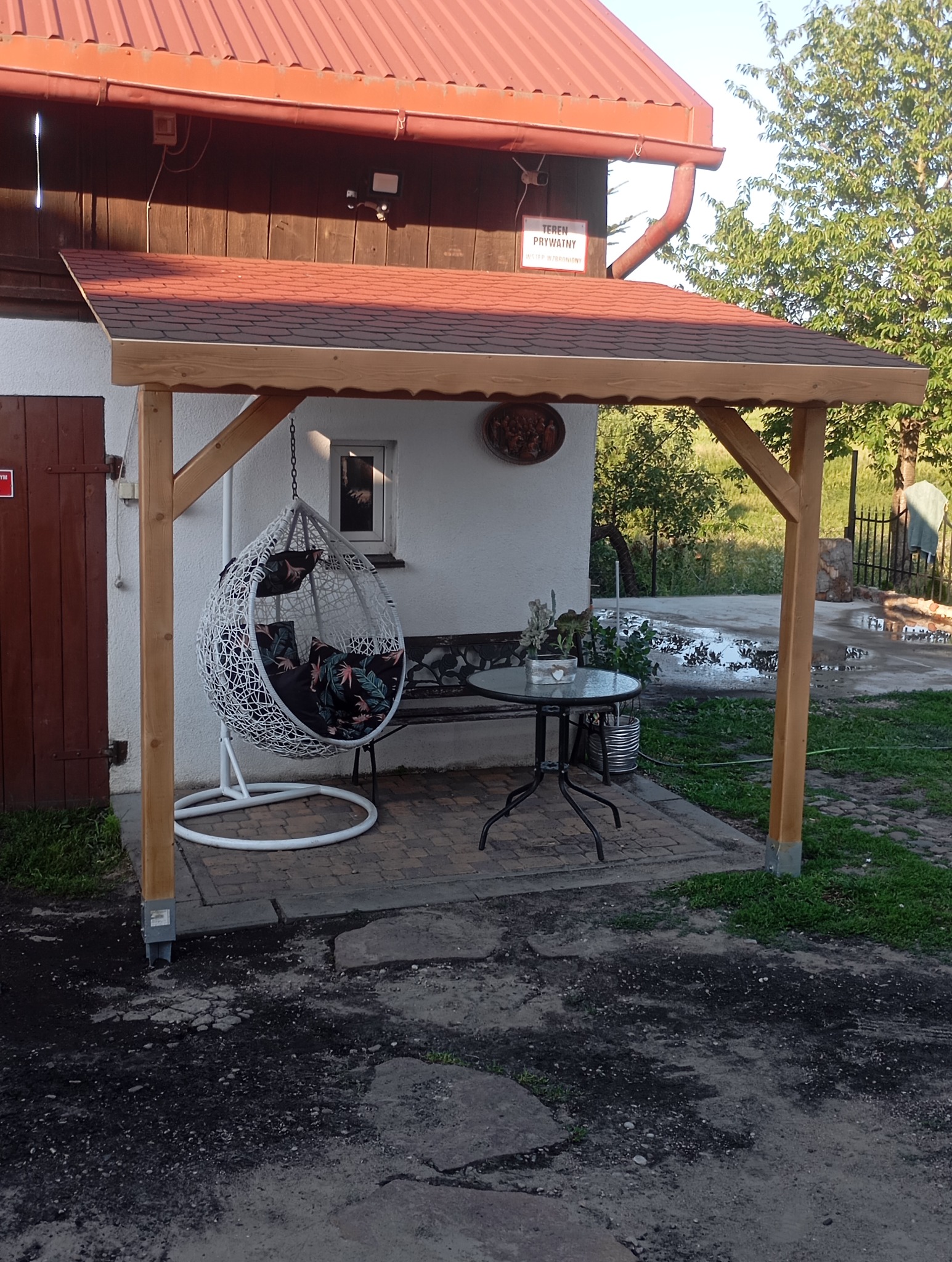 Drewniana pergola z czerwoną dachówką, pod nią okrągły stolik, wiszące krzesło w stylu boho i ławka na tle białej ściany budynku z brązowymi drzwiami i małym oknem.