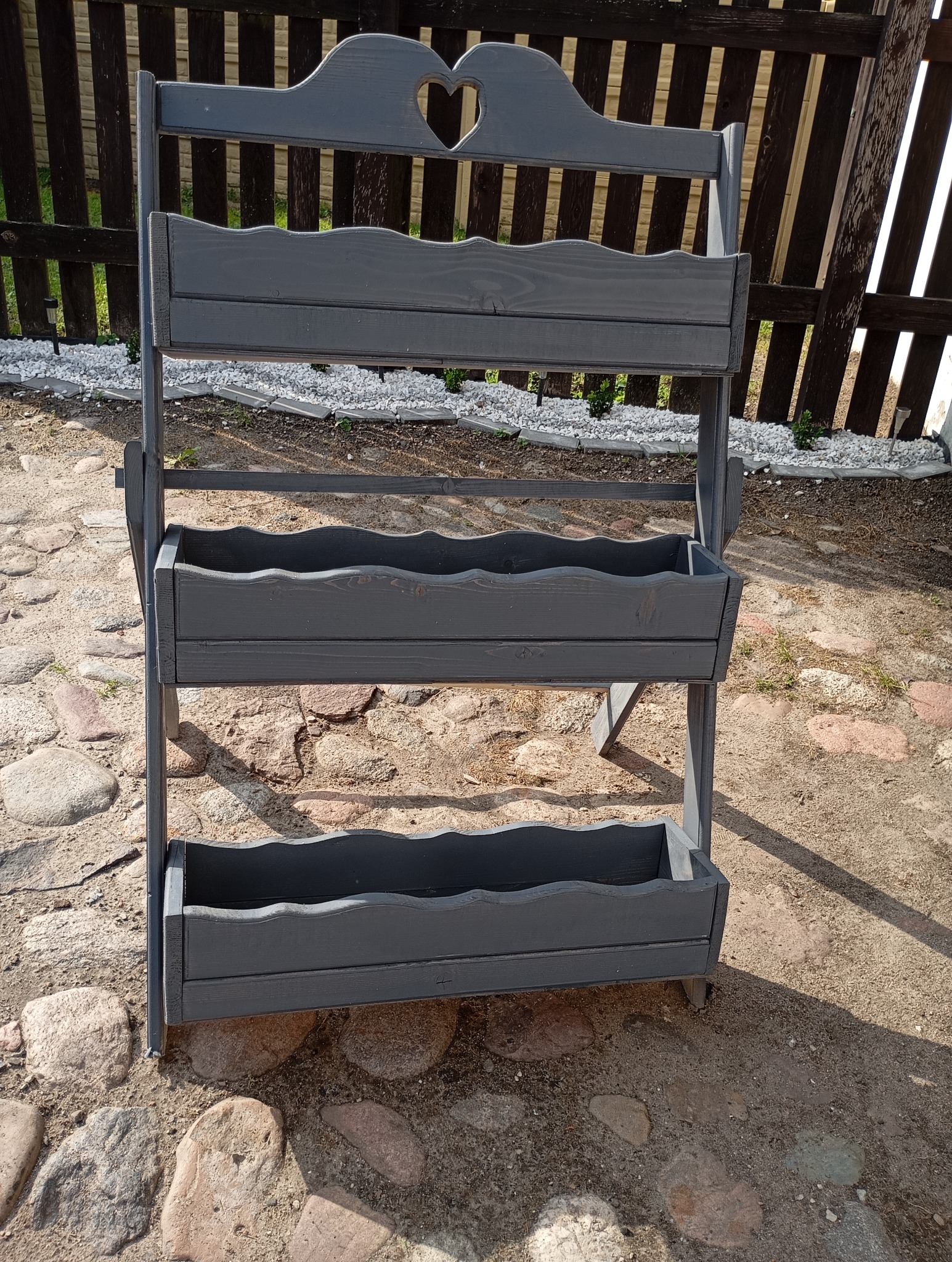 Trzypoziomowy, szary, drewniany kwietnik z dekoracyjnym wycięciem w kształcie serca w górnej części, stojący na kamiennej nawierzchni przed drewnianym płotem.