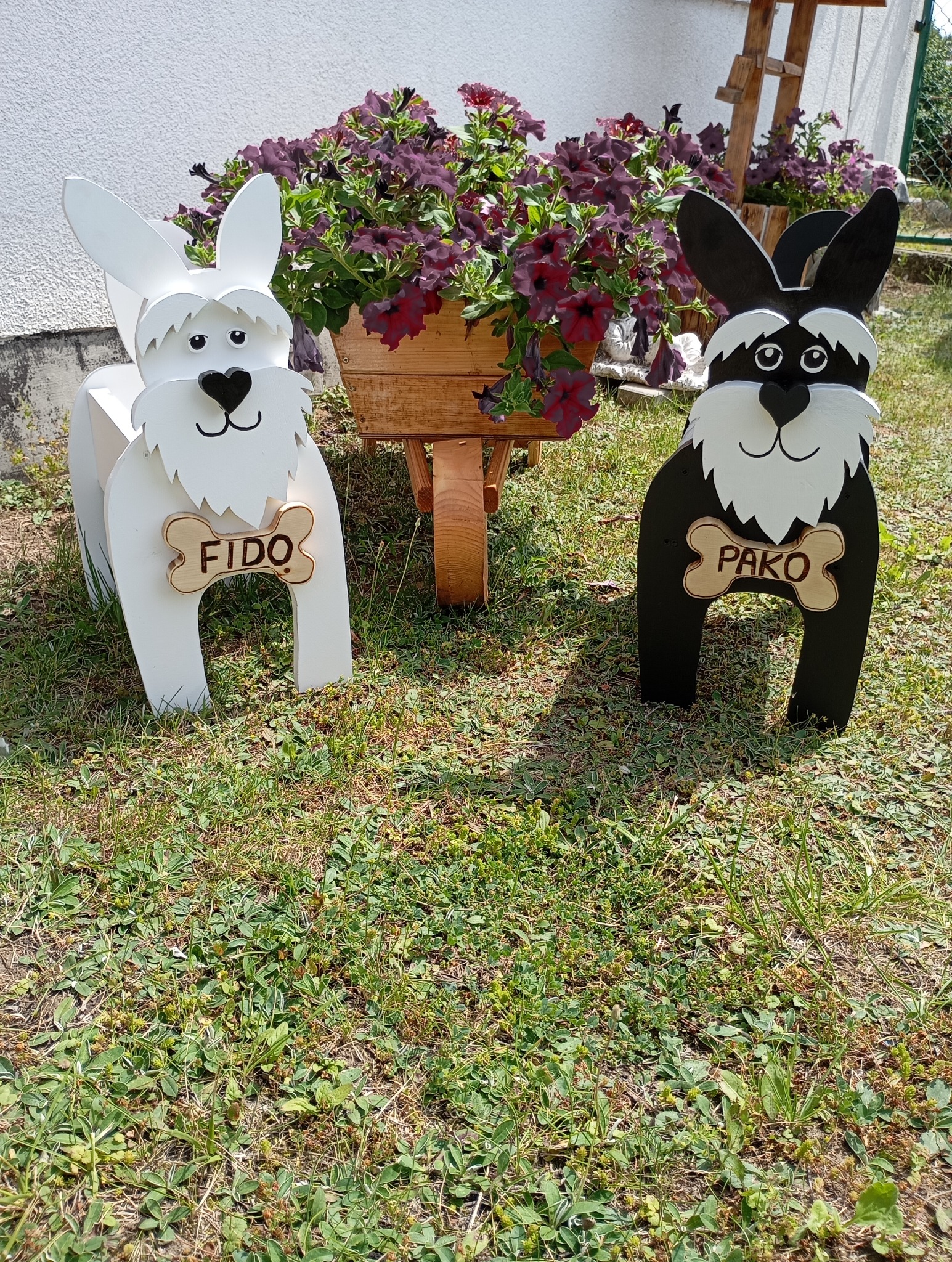 Dwie drewniane dekoracje ogrodowe w kształcie psów, biała z imieniem Fido i czarna z imieniem Pako, stojące na trawie obok drewnianej taczki z fioletowymi kwiatami.