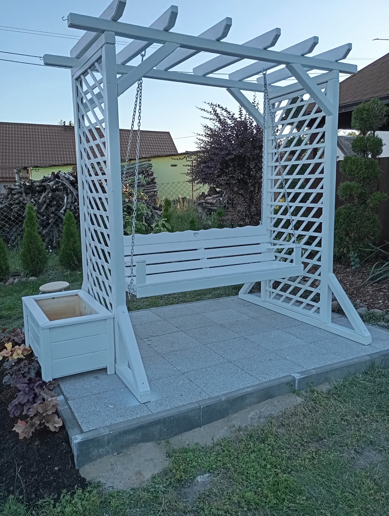 Biała drewniana pergola z huśtawką, kratownicowe ściany boczne, donica z boku, ułożona na szarych płytach chodnikowych, w tle dom i zieleń.