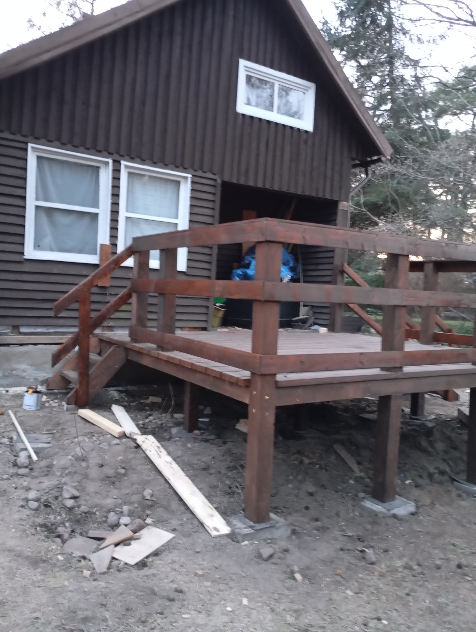 Drewniany taras w trakcie budowy przy brązowym domu z białymi oknami, widoczne deski, podpory i narzędzia na ziemi.