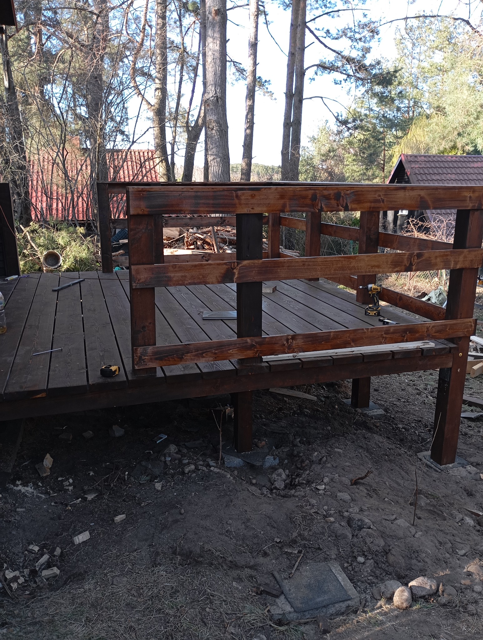 Budowa ciemnobrązowego tarasu z drewnianą balustradą, widoczne narzędzia i drzewa w tle.