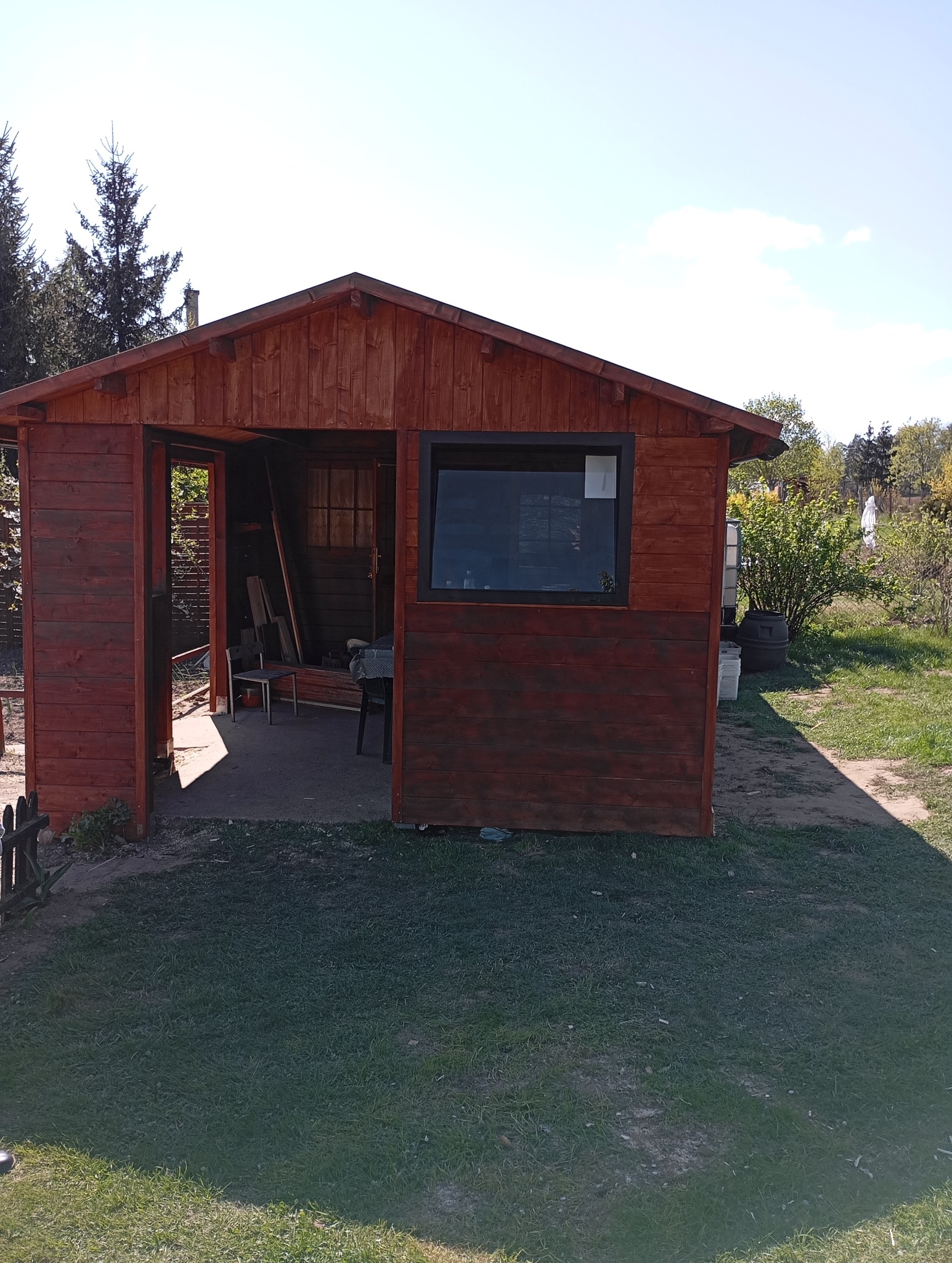 Drewniana altana ogrodowa z oknem i otwartymi drzwiami, pomalowana na brązowo, otoczona zielenią. Widoczne elementy wyposażenia wewnątrz.