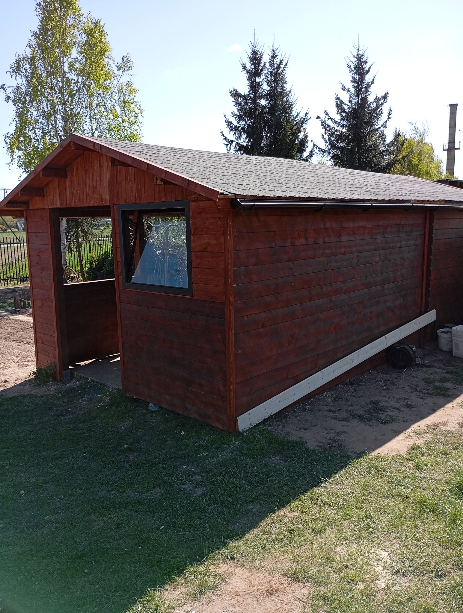 Ciemnobrązowa drewniana altana z oknem i otwartą przestrzenią, widoczna na trawniku. Elementy konstrukcyjne w trakcie montażu. W tle drzewa i błękitne niebo.