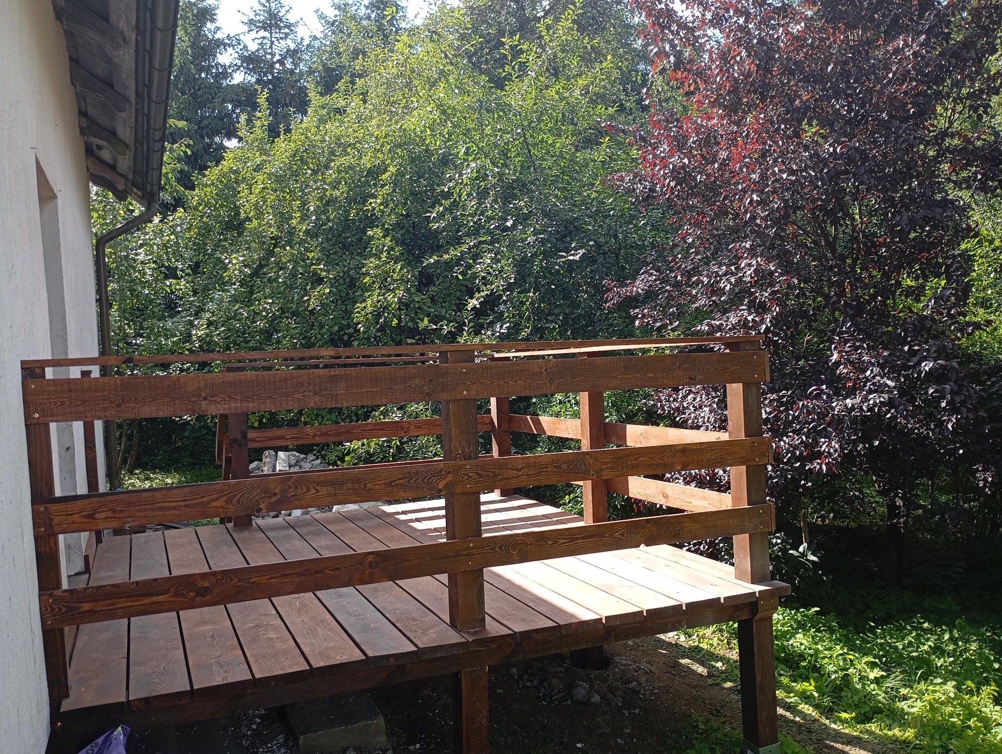 Świeżo wykończony, drewniany taras z bejcowaną balustradą, przylegający do budynku. W tle bujna roślinność i drzewo o purpurowych liściach. Słoneczny dzień.