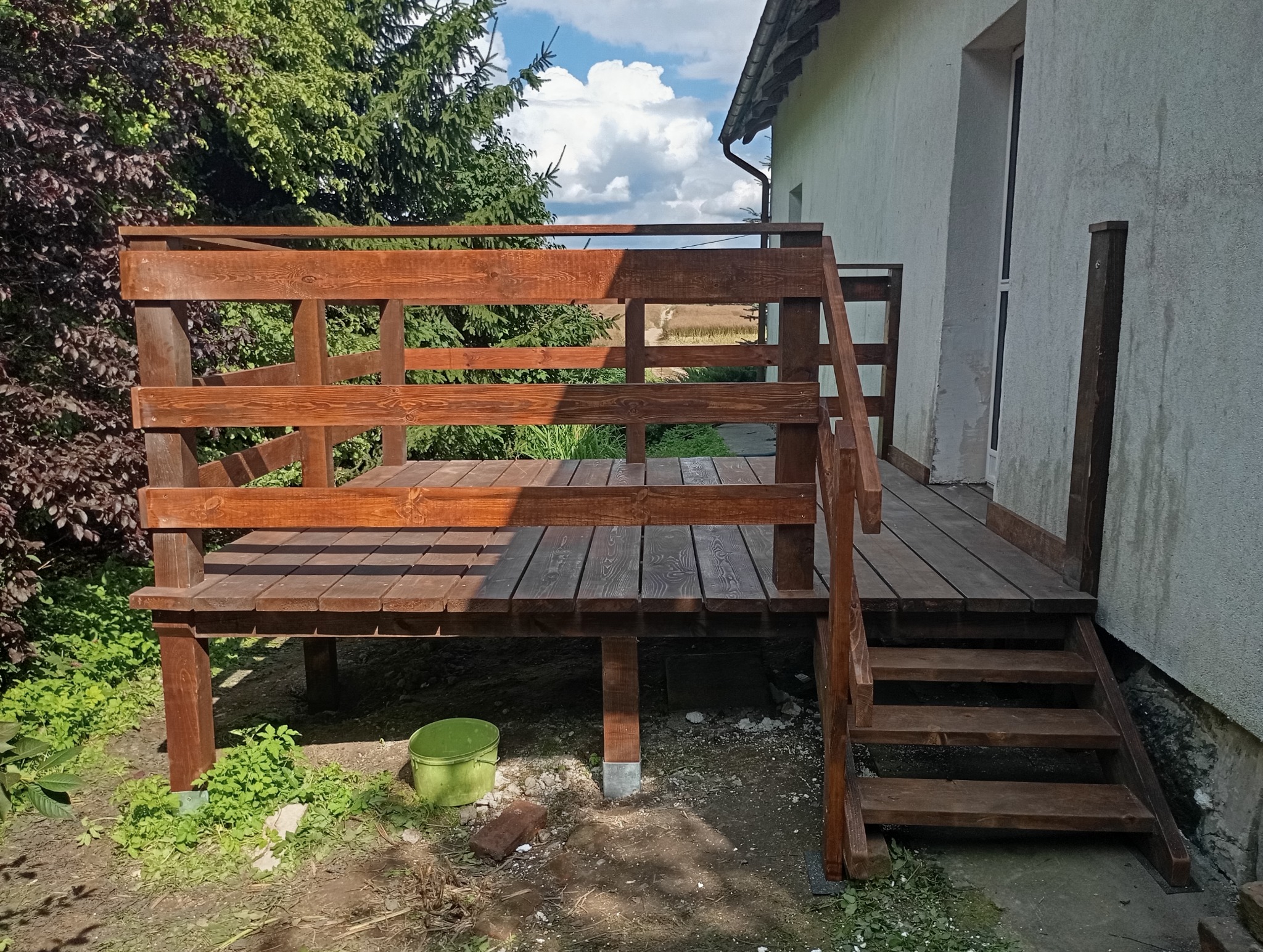 Drewniany taras z balustradą i schodami, świeżo po impregnacji, przylegający do jasnego budynku. Widok z dołu na tle zieleni i błękitnego nieba.