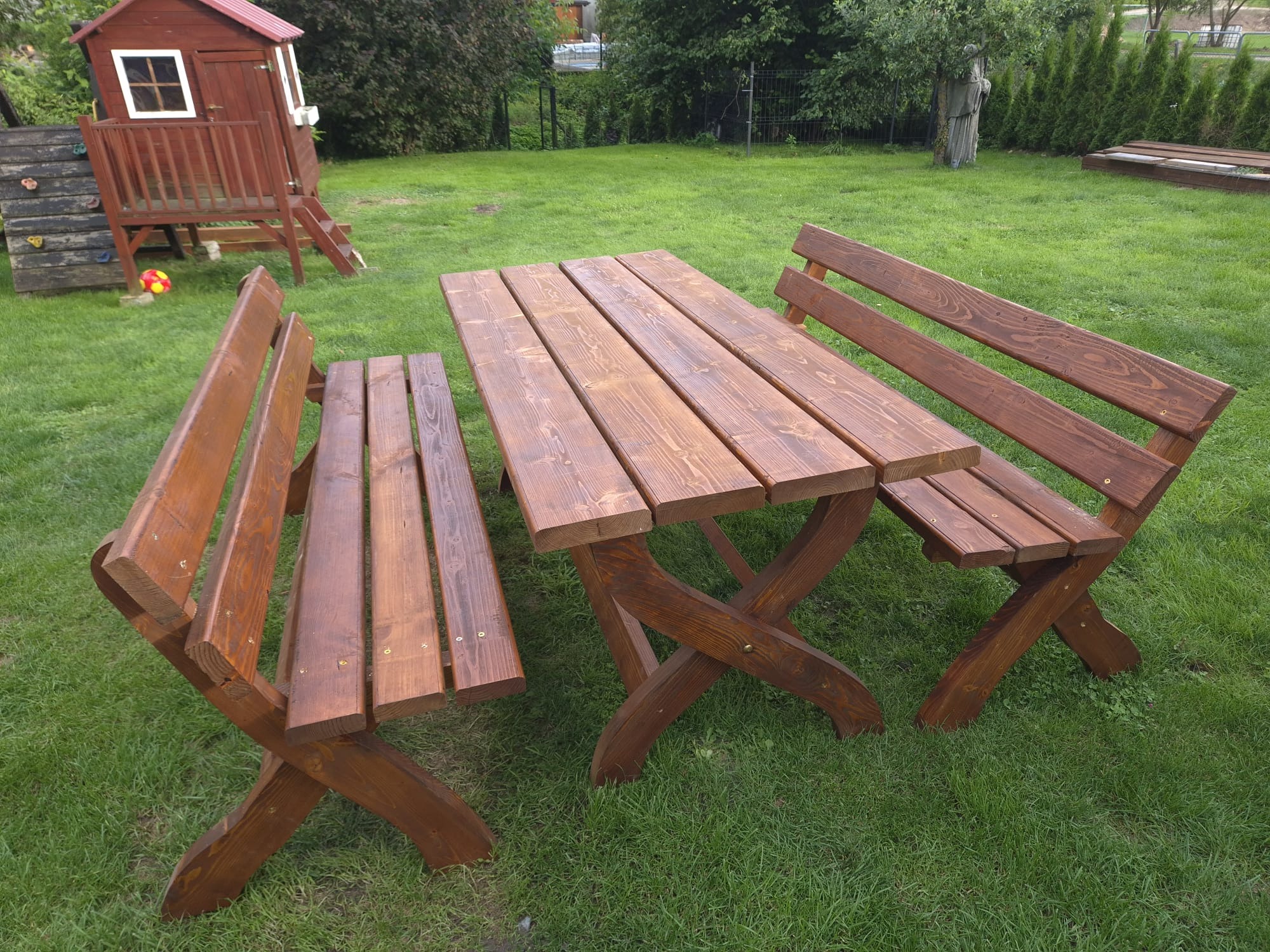 Drewniany stół piknikowy z dwiema ławkami na trawniku, w tle domek dla dzieci i drzewa. Widoczne detale konstrukcyjne i faktura drewna.