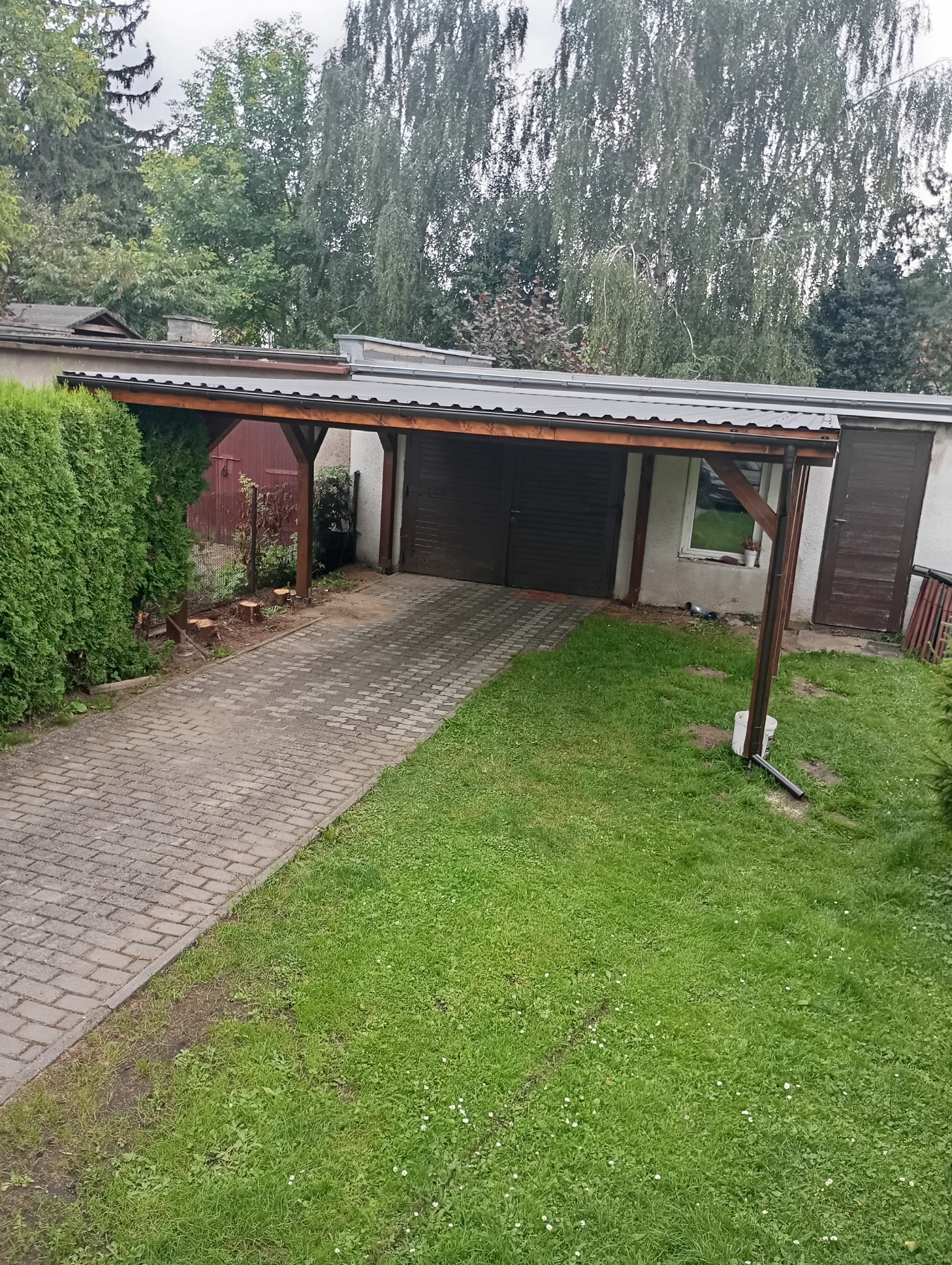 Zadaszenie nad podjazdem z drewnianą konstrukcją, widok z perspektywy ogrodu, z kostką brukową i zielenią. Fragment budynku z bramą garażową w tle.