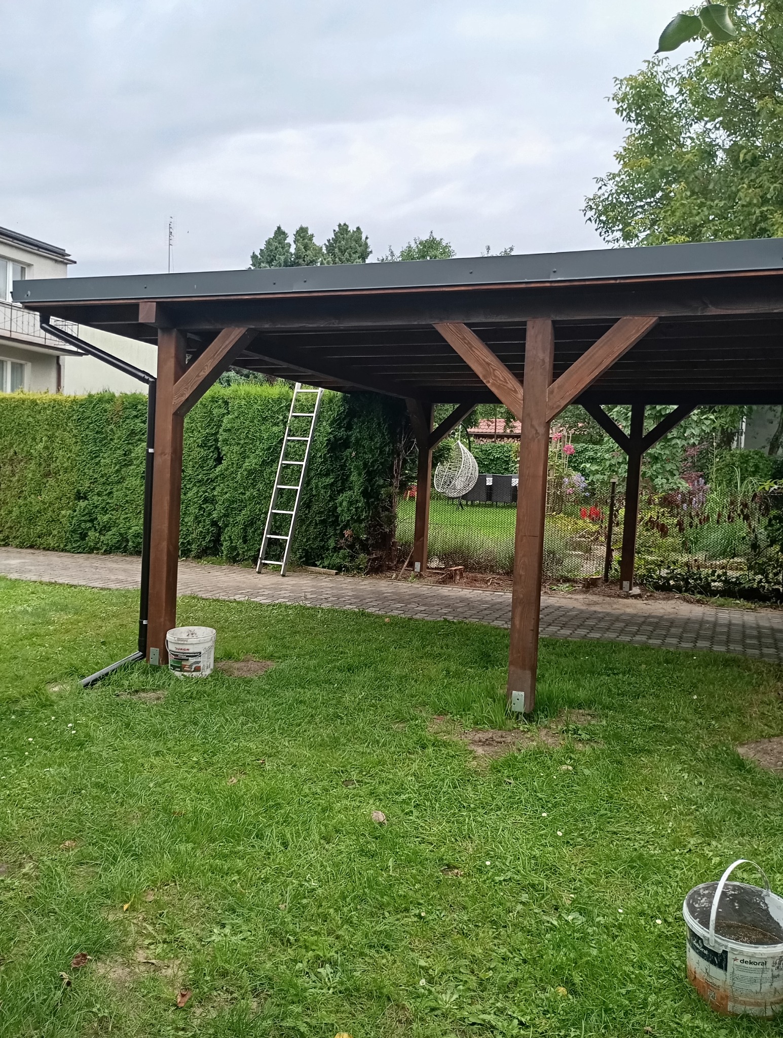 Drewniana pergola z ciemnym dachem w ogrodzie, widoczna drabina i wiadro z farbą. Ujęcie z poziomu trawy, na tle zieleni i części domu.