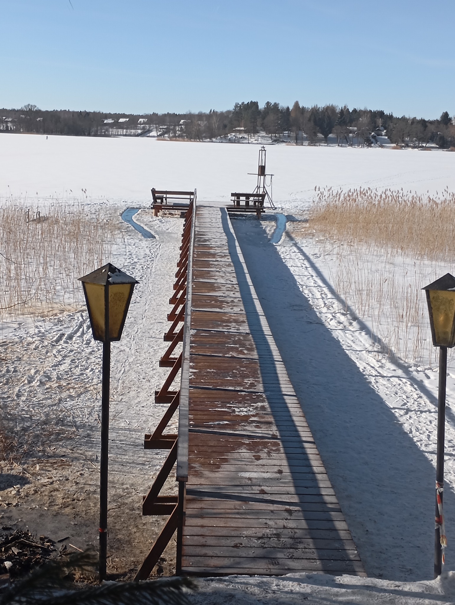 Zimowy krajobraz z drewnianym pomostem prowadzącym przez ośnieżone jezioro. Dwie ławki na końcu pomostu. Po bokach pomostu lampy oświetleniowe.