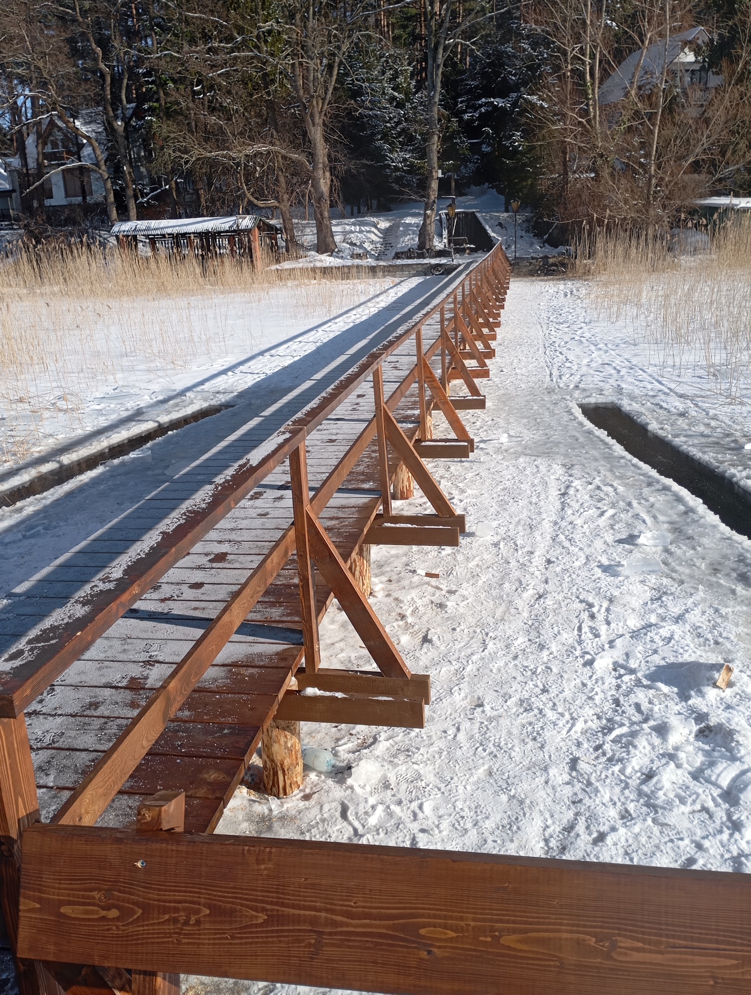 Pomost z drewnianą balustradą prowadzący przez zasypany śniegiem teren. Widoczne ślady na śniegu i częściowo zamarznięte jezioro. Słoneczny dzień.