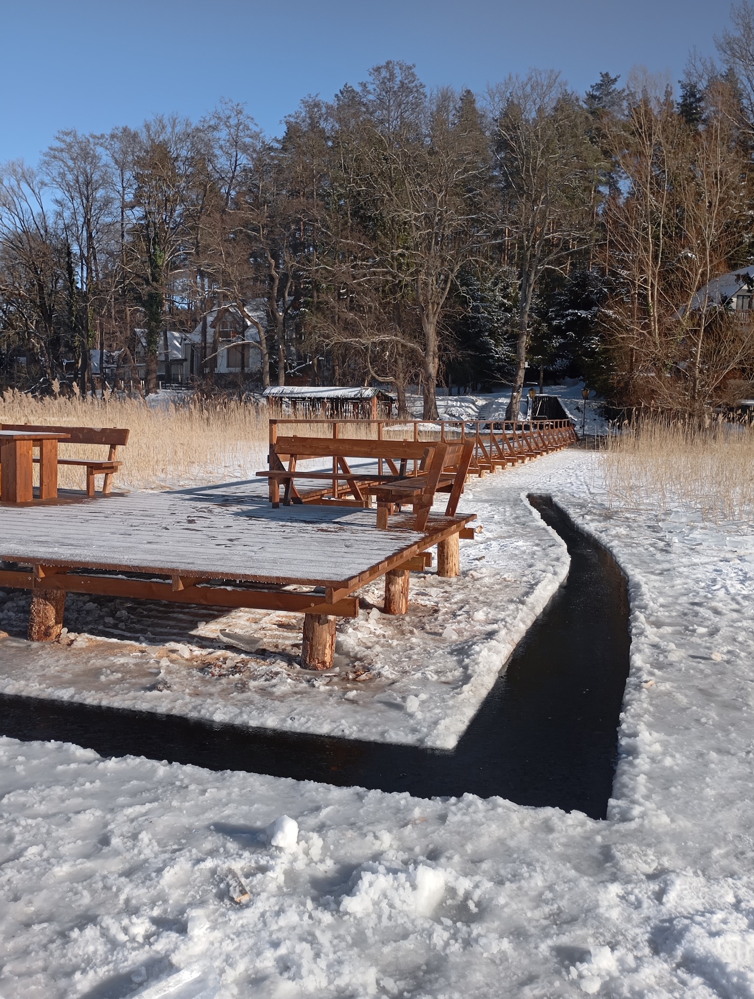 Zimowy krajobraz z drewnianym pomostem i ławkami nad zamarzniętym jeziorem. Śnieg pokrywa konstrukcję i otoczenie, tworząc malowniczy widok.