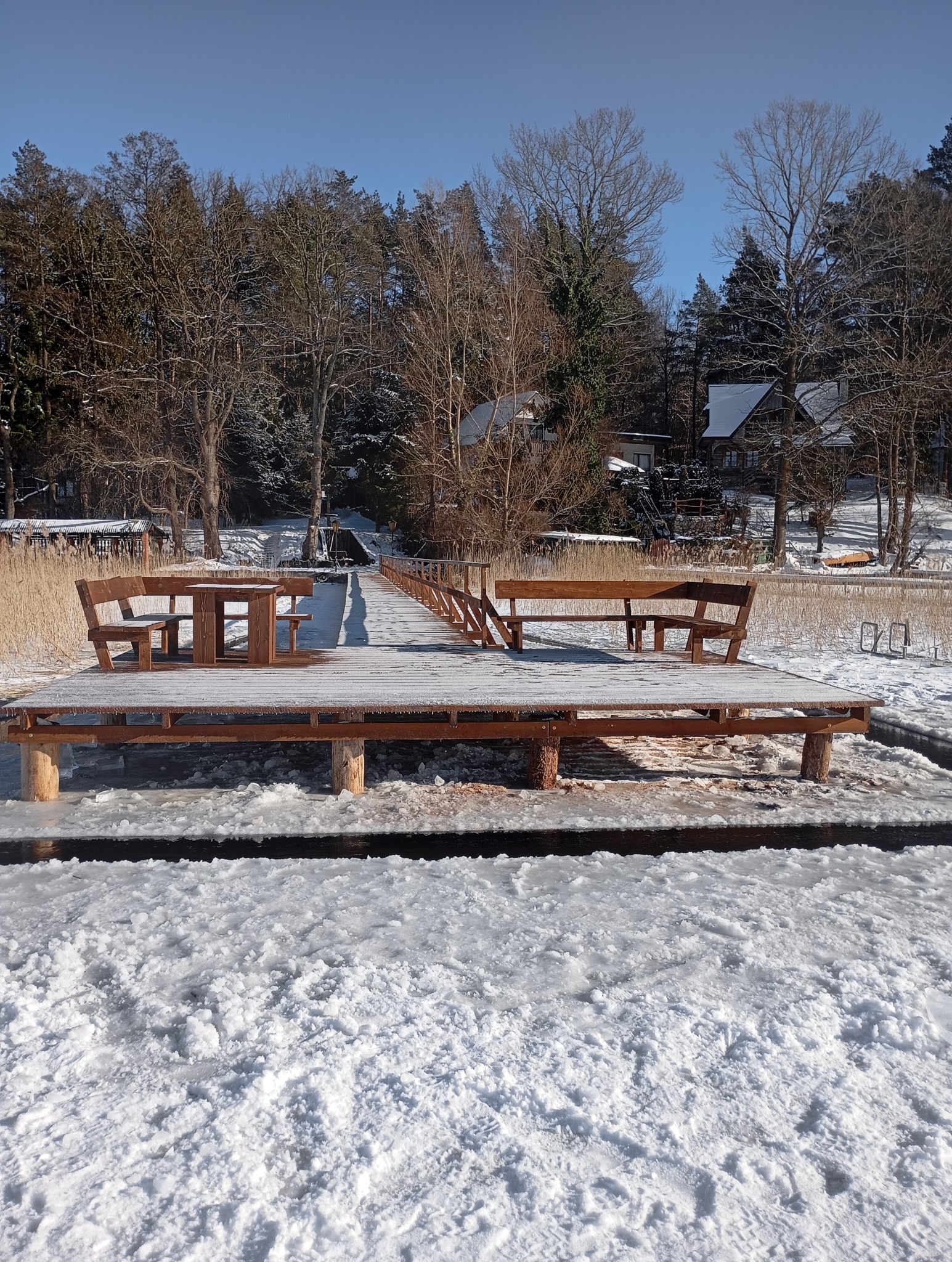 Zimowy krajobraz z drewnianym pomostem nad zamarzniętym jeziorem. Na pomoście ustawiony stół i ławki. W tle las i domy.