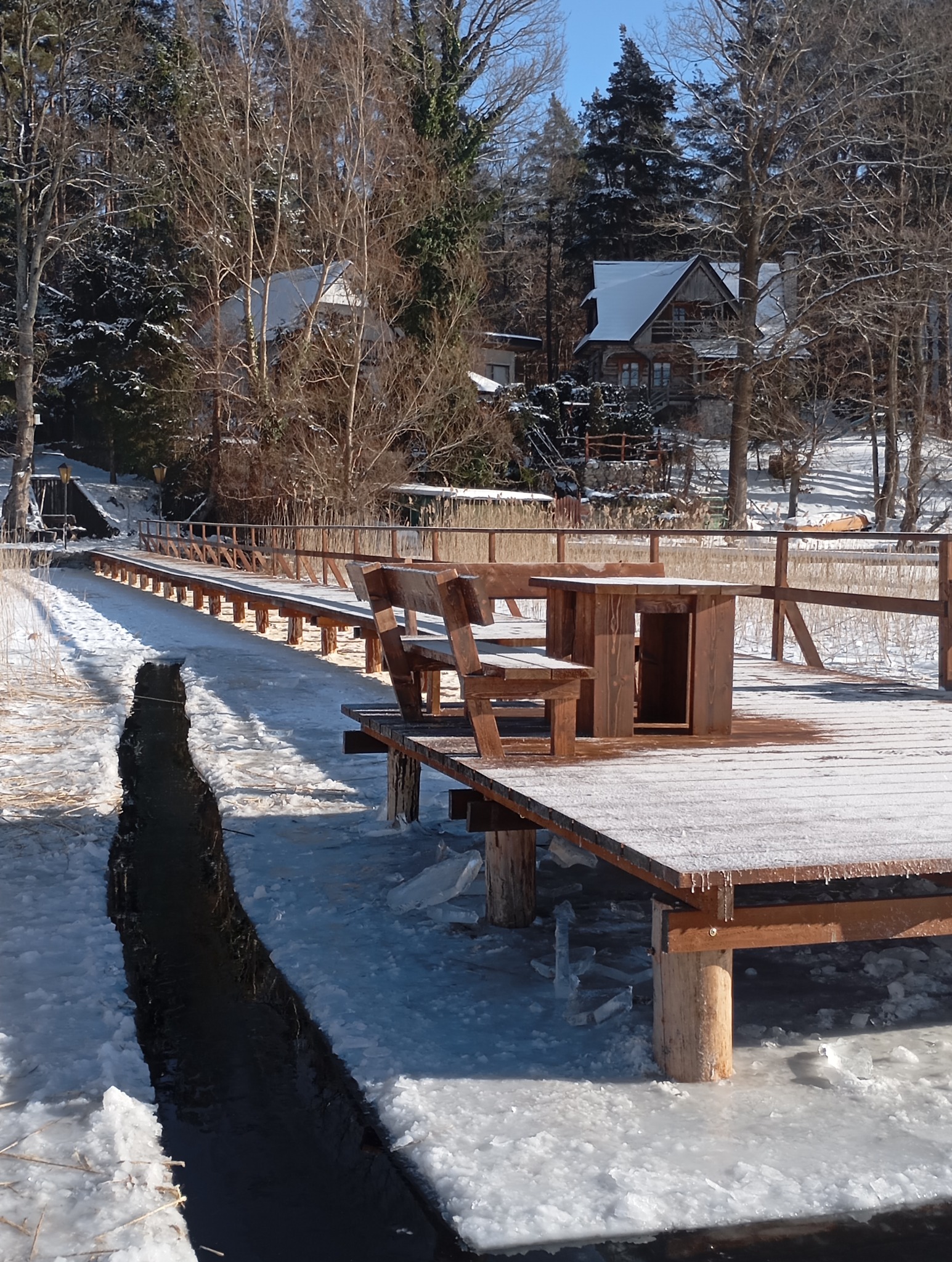 Zimowy krajobraz z drewnianym tarasem nad wodą, na którym stoi stół i ławka. W tle ośnieżone drzewa i dom. Całość pokryta szronem.
