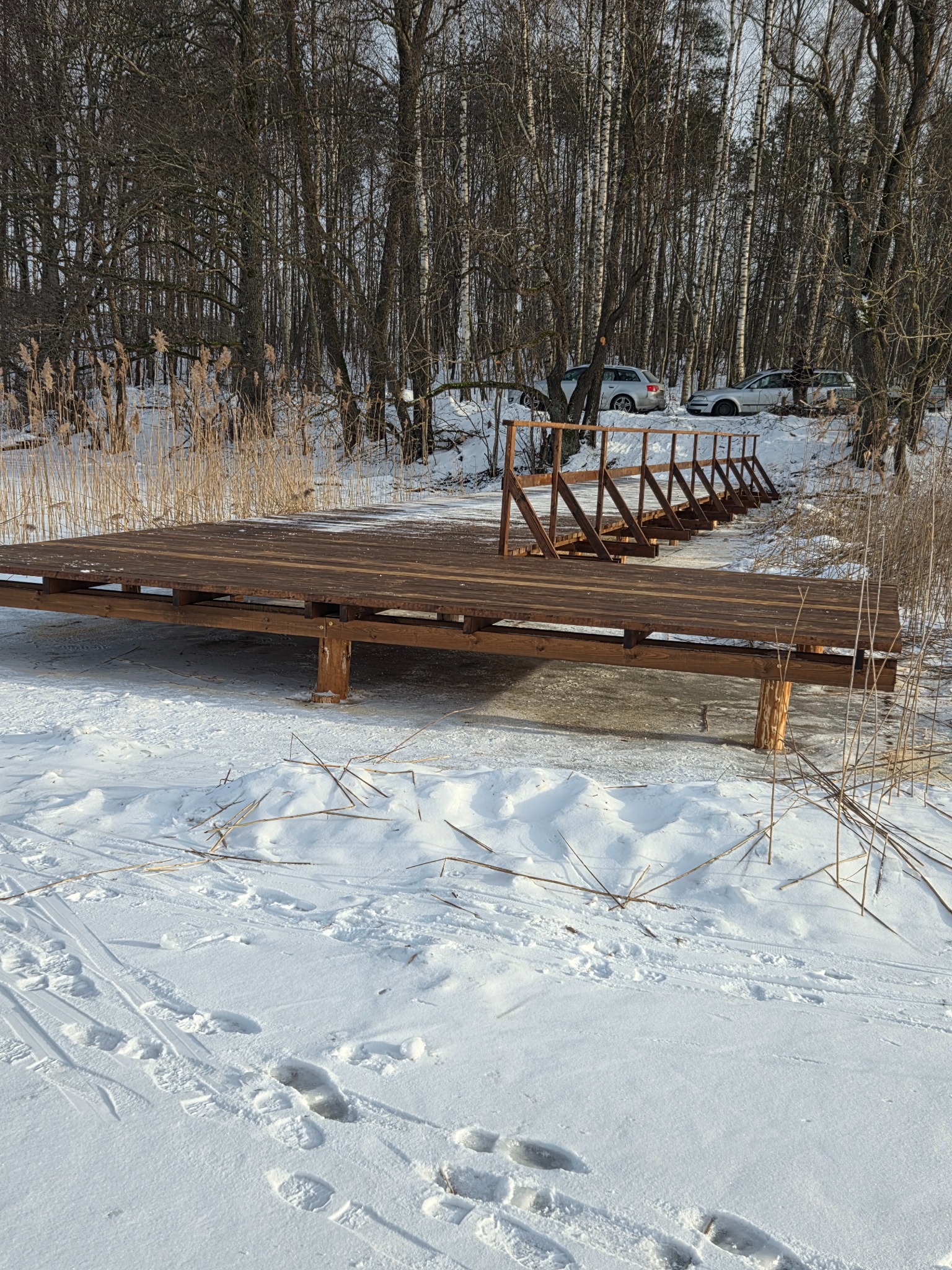 Pomost z drewnianą balustradą na zamarzniętym jeziorze, otoczony trzcinami i lasem. W tle zaparkowane samochody. Zimowy krajobraz.