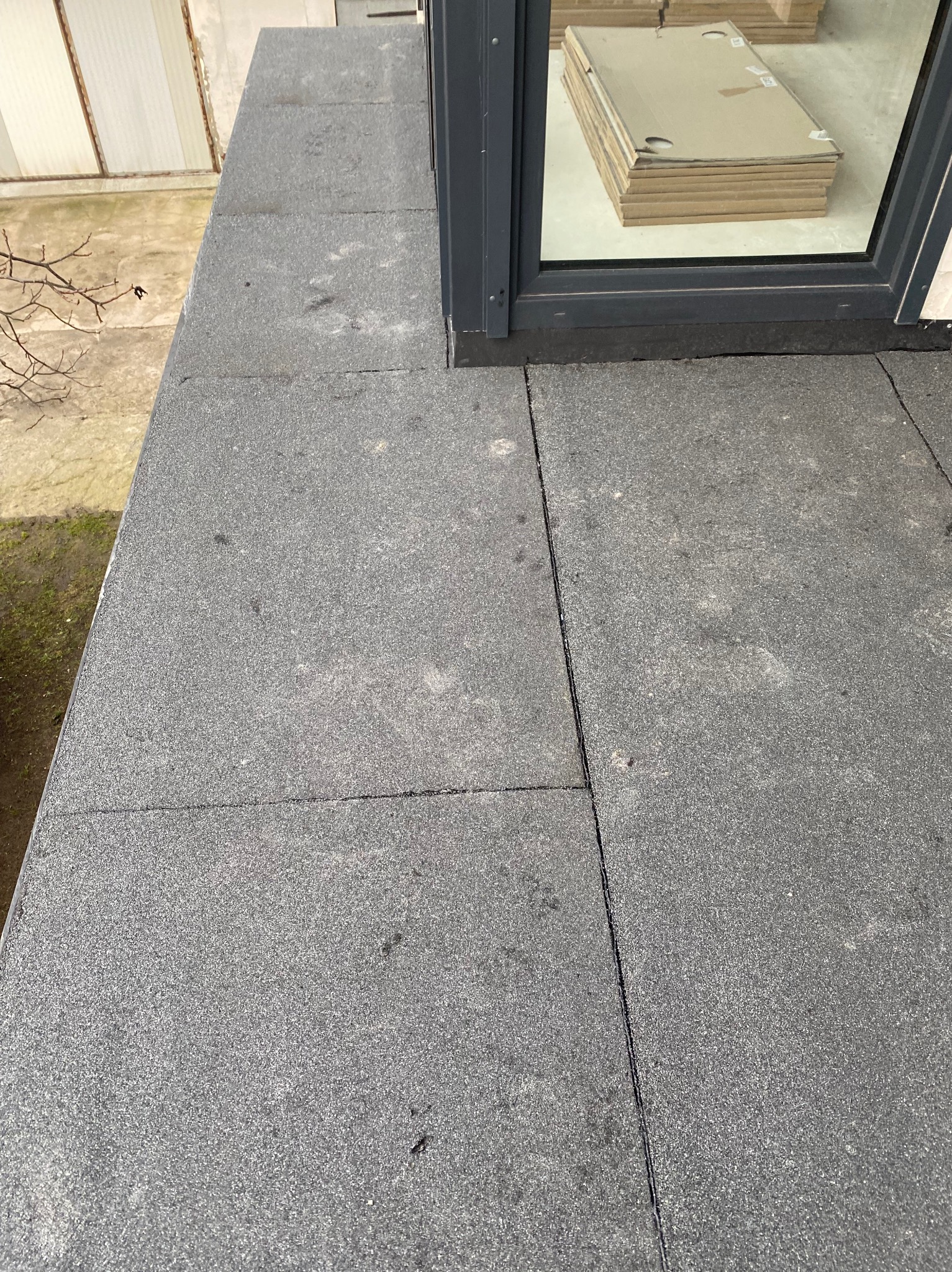 Szara, gumowa powierzchnia dachu z widocznym oknem dachowym i stosem materiałów budowlanych za szybą.