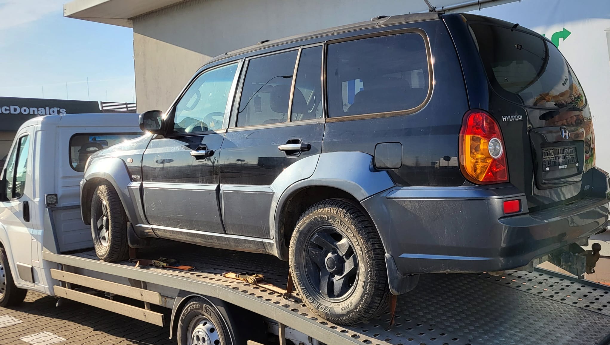 Czarny samochód Hyundai Terracan na platformie białej lawety, widok z boku, w tle budynek z logo McDonald's.