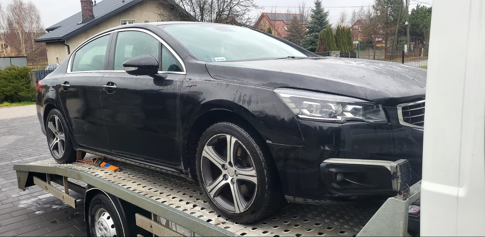 Czarny samochód osobowy marki Peugeot z widocznym znaczkiem GT na błotniku, zabezpieczony pasami na platformie lawety, na tle zabudowań mieszkalnych w pochmurny dzień.