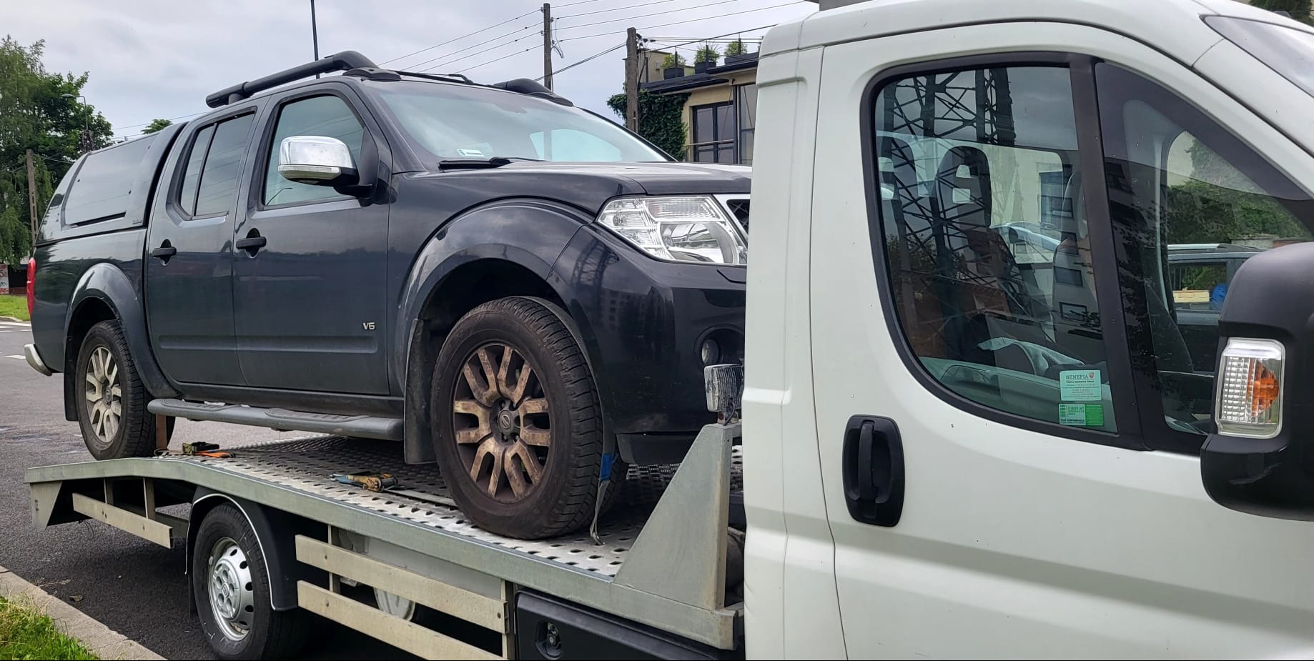 Czarny Nissan Navara V6 z zabudową na pace, z brudnymi felgami, załadowany na lawetę, widoczna kabina białego samochodu lawety z naklejkami na szybie.