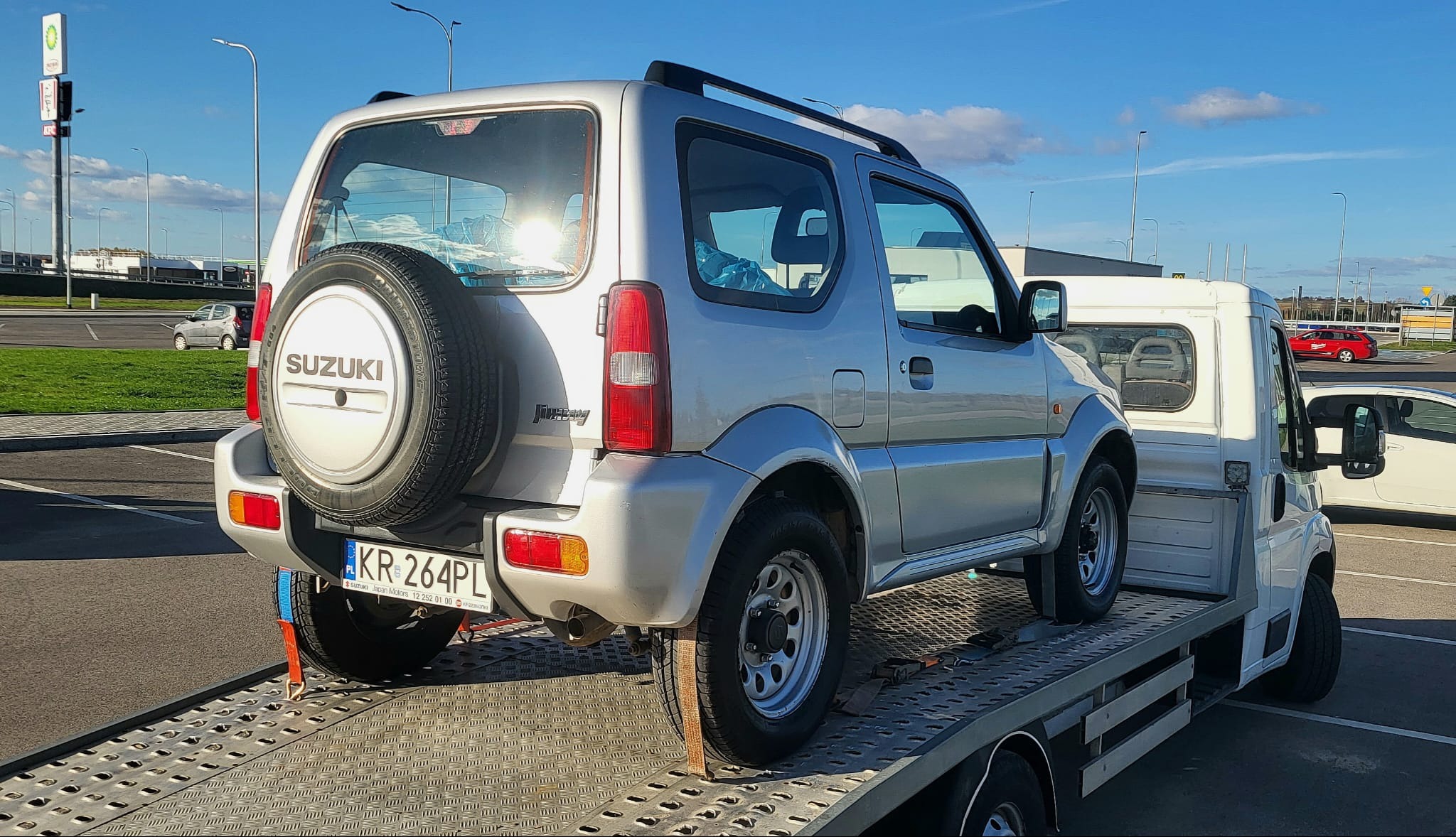 Srebrny Suzuki Jimny zabezpieczony pasami na platformie lawety, widok z tyłu, tablica rejestracyjna KR 264PL, w tle parking i słup z logo BP.