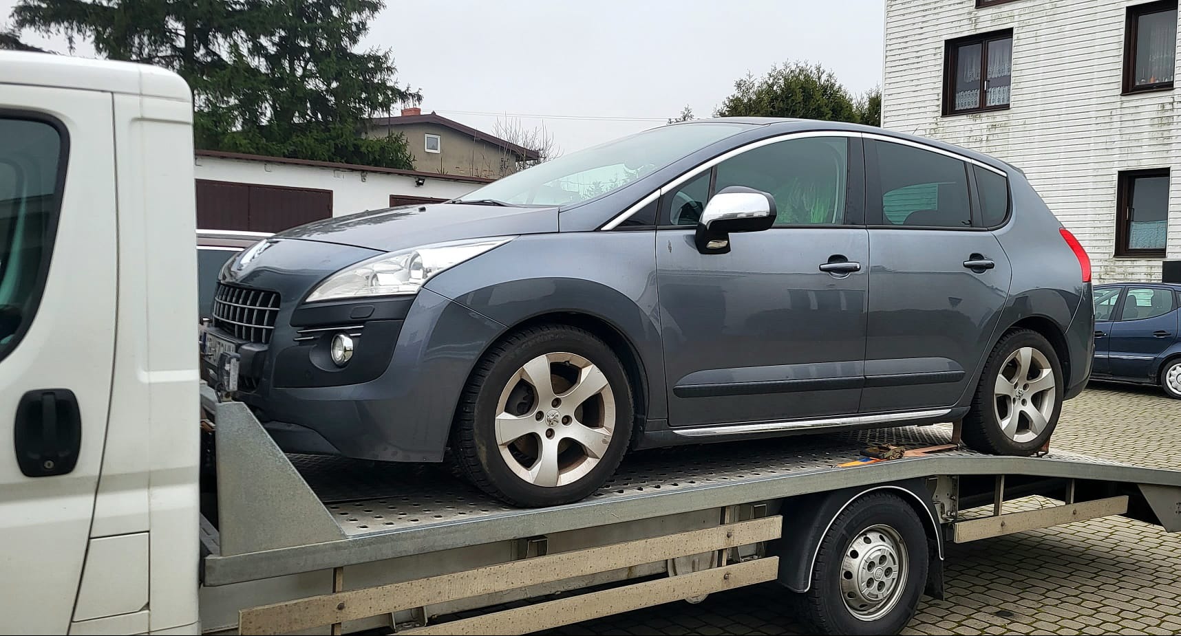 Szary samochód Peugeot 3008 na platformie lawety, zabezpieczony pasami transportowymi, widoczny z boku na tle budynku z szarą elewacją.