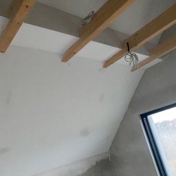 Nikon Construction Heorhii Nikonenko - Skośny sufit poddasza z drewnianą konstrukcją i częściowo położoną płytą gipsową, widok z dołu, Kraków. Widoczne kable elektryczne.