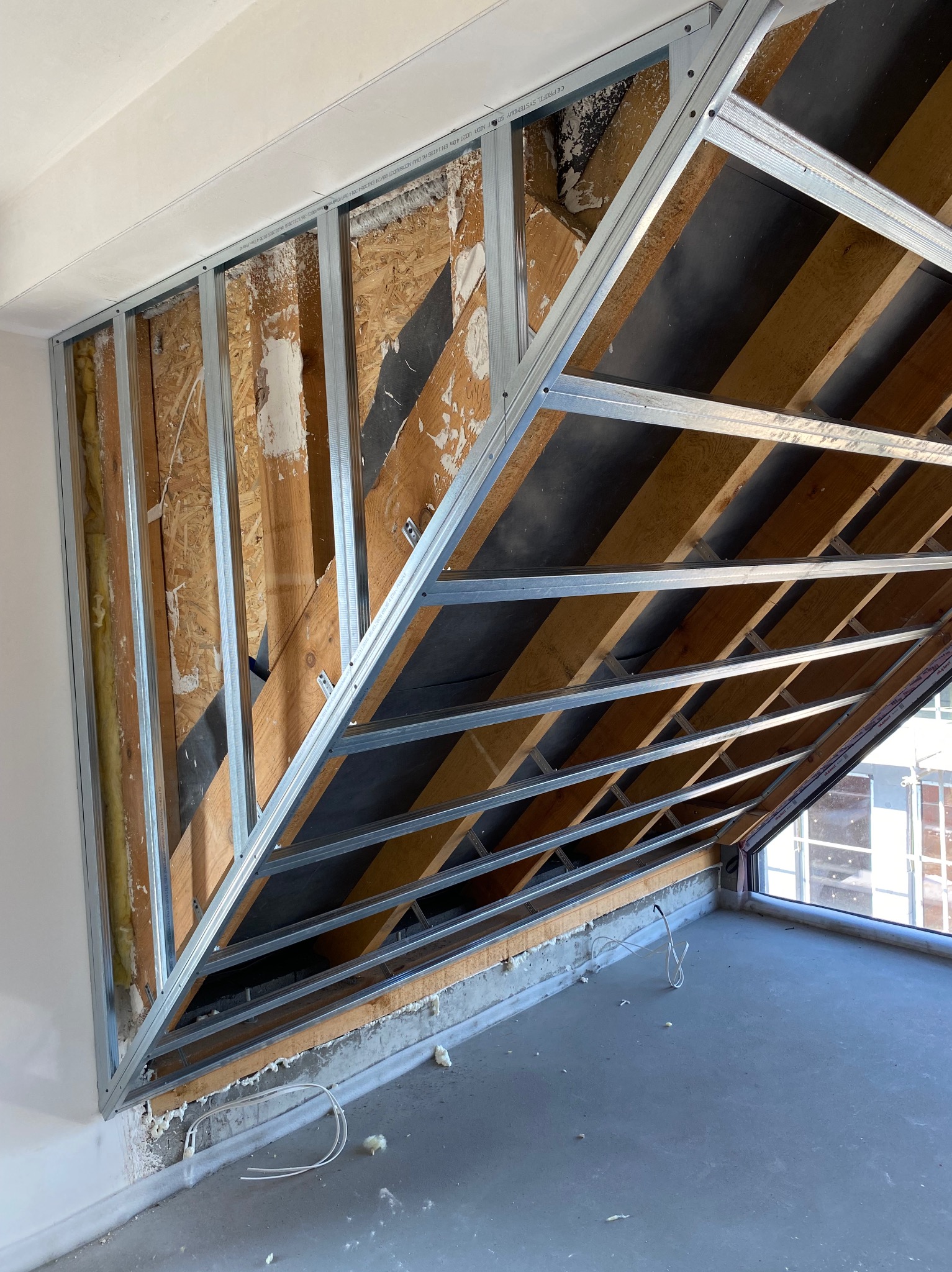 Szkielet metalowy pod zabudowę poddasza z widoczną izolacją i elementami drewnianymi, przygotowany do dalszych prac wykończeniowych. Widok na okno dachowe.