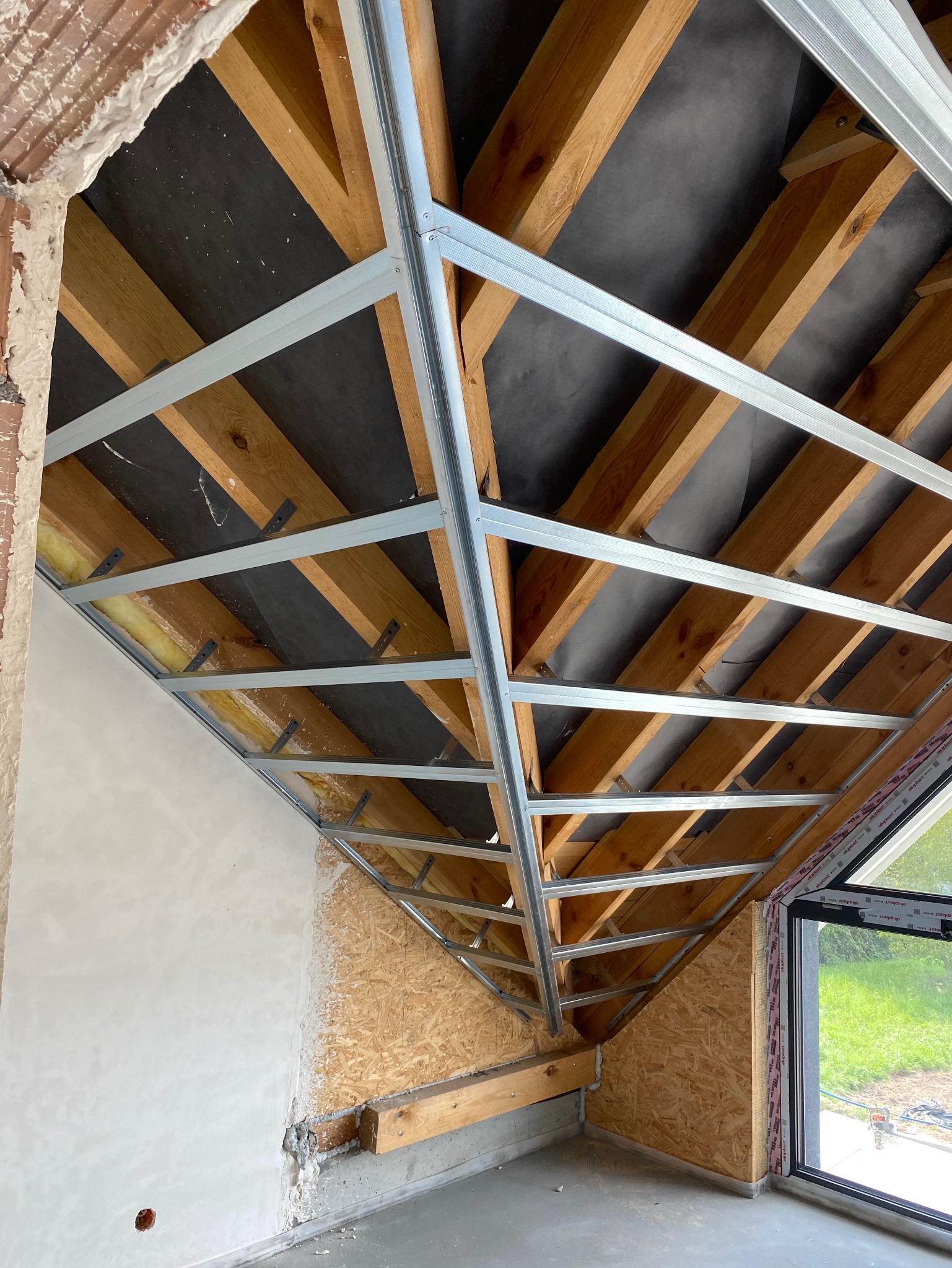 Skośny sufit poddasza w trakcie montażu stelaża z metalowych profili, widoczne drewniane krokwie i czarna membrana. Ściana z fragmentami tynku i płyty OSB.