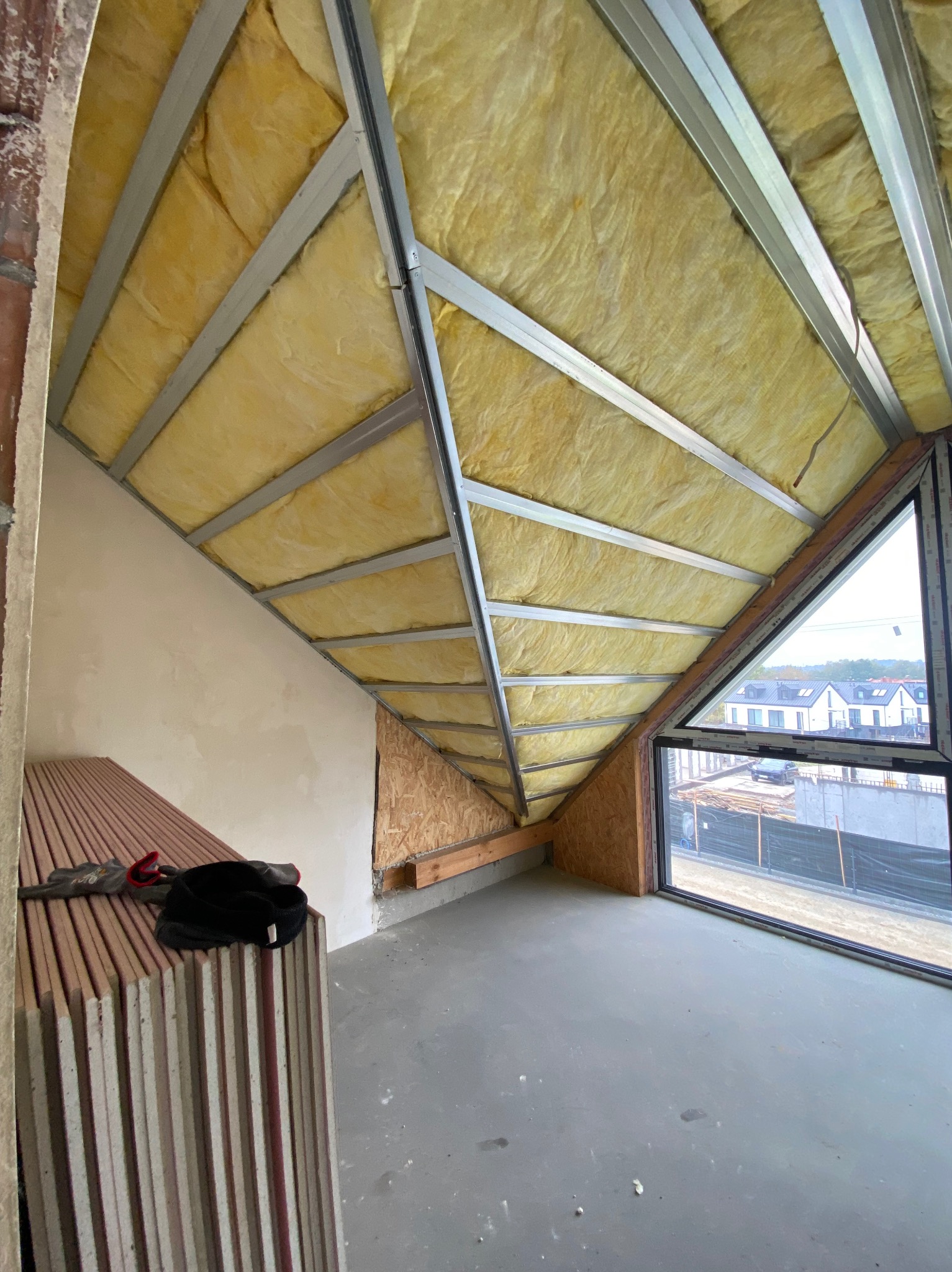 Poddasze z oknem w trakcie ocieplania: wełna mineralna między metalowymi profilami, przygotowane płyty gipsowe, surowy beton na podłodze. Widok na budynki.
