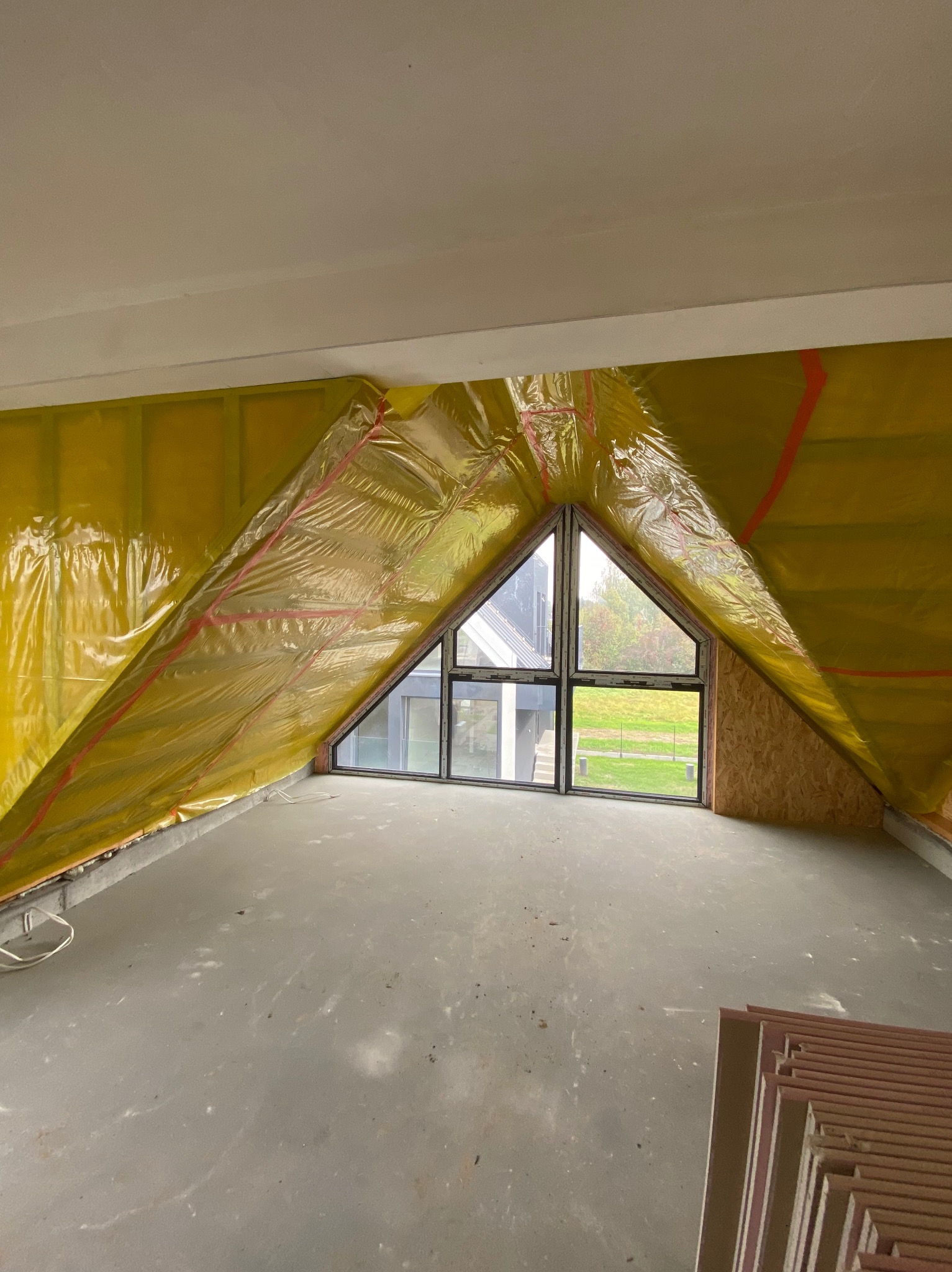 Poddasze z oknem trójkątnym, ocieplone żółtą folią z czerwonymi paskami, widoczne płyty izolacyjne. Etap prac przed wykończeniem.