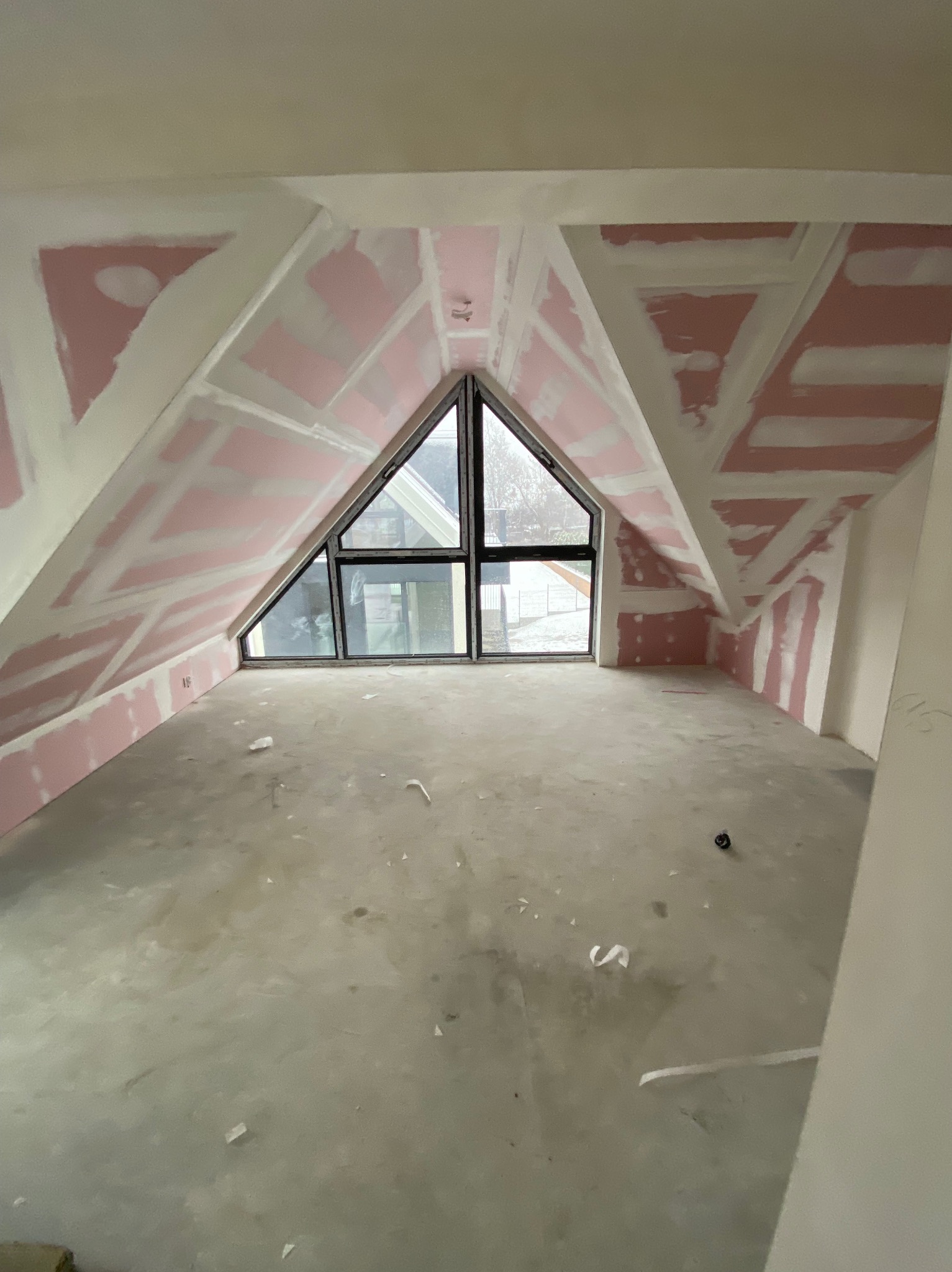 Poddasze z oknem w kształcie trójkąta, ściany wykończone płytami kartonowo-gipsowymi, przygotowane do dalszych prac wykończeniowych. Widoczne łączenia płyt.