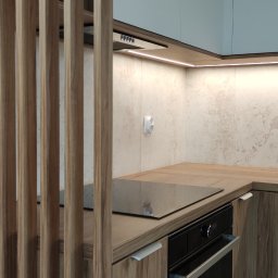 PRZESTRZEŃ SPOTKANIA Mariusz Kaczmarczyk - Fragment kuchni z drewnianymi szafkami, wbudowanym piekarnikiem i płytą indukcyjną, oddzielony ażurową ścianką z drewnianych listew.