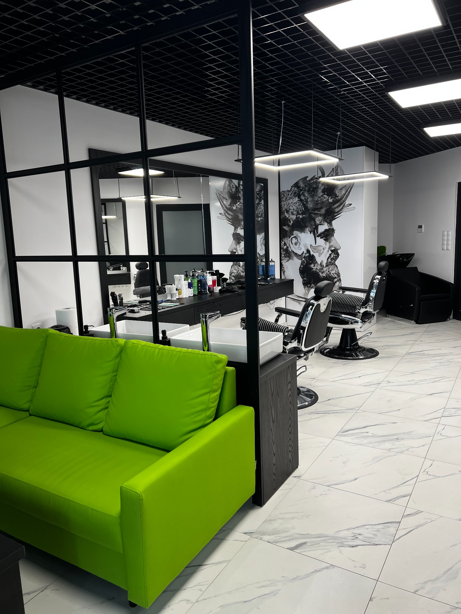 Nowoczesne wnętrze salonu fryzjerskiego z zieloną sofą, stanowiskami z umywalkami, fryzjerskimi fotelami i czarno-białym portretem na ścianie.