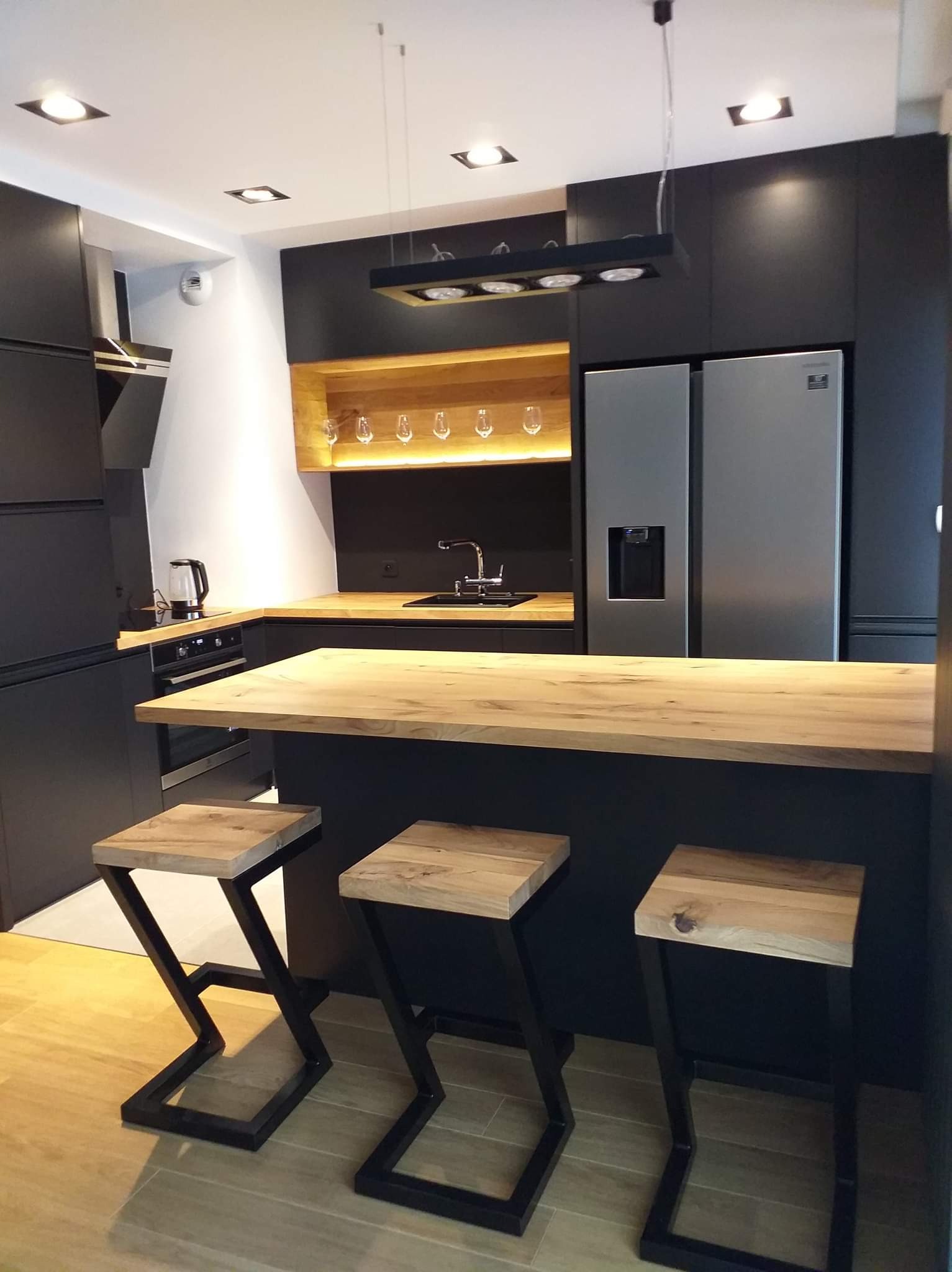 Nowoczesna kuchnia w czerni i drewnie z wyspą i trzema hokerami z metalowymi nogami w kształcie litery Z, oświetlenie punktowe i półka na wino.