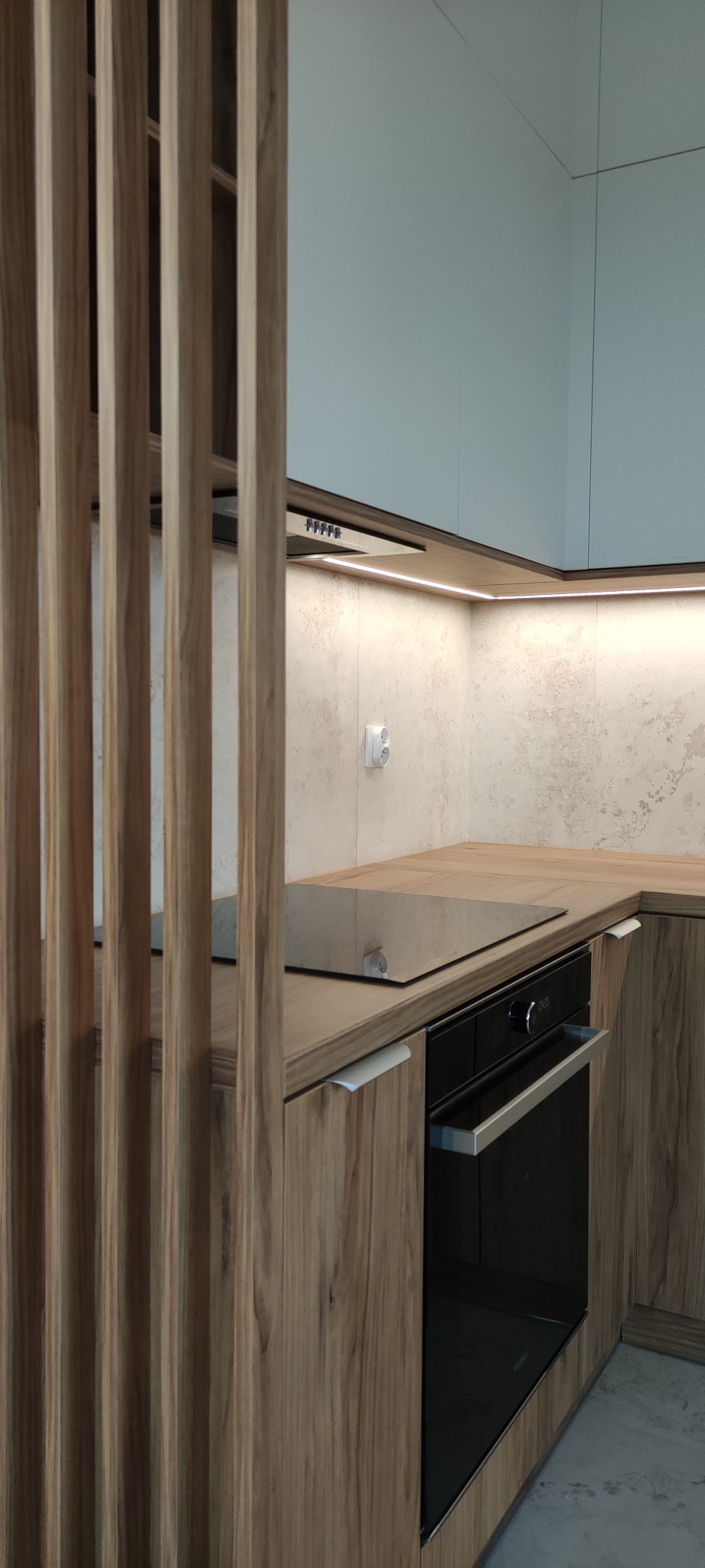 Fragment kuchni z drewnianymi szafkami, wbudowanym piekarnikiem i płytą indukcyjną, oddzielony ażurową ścianką z drewnianych listew.