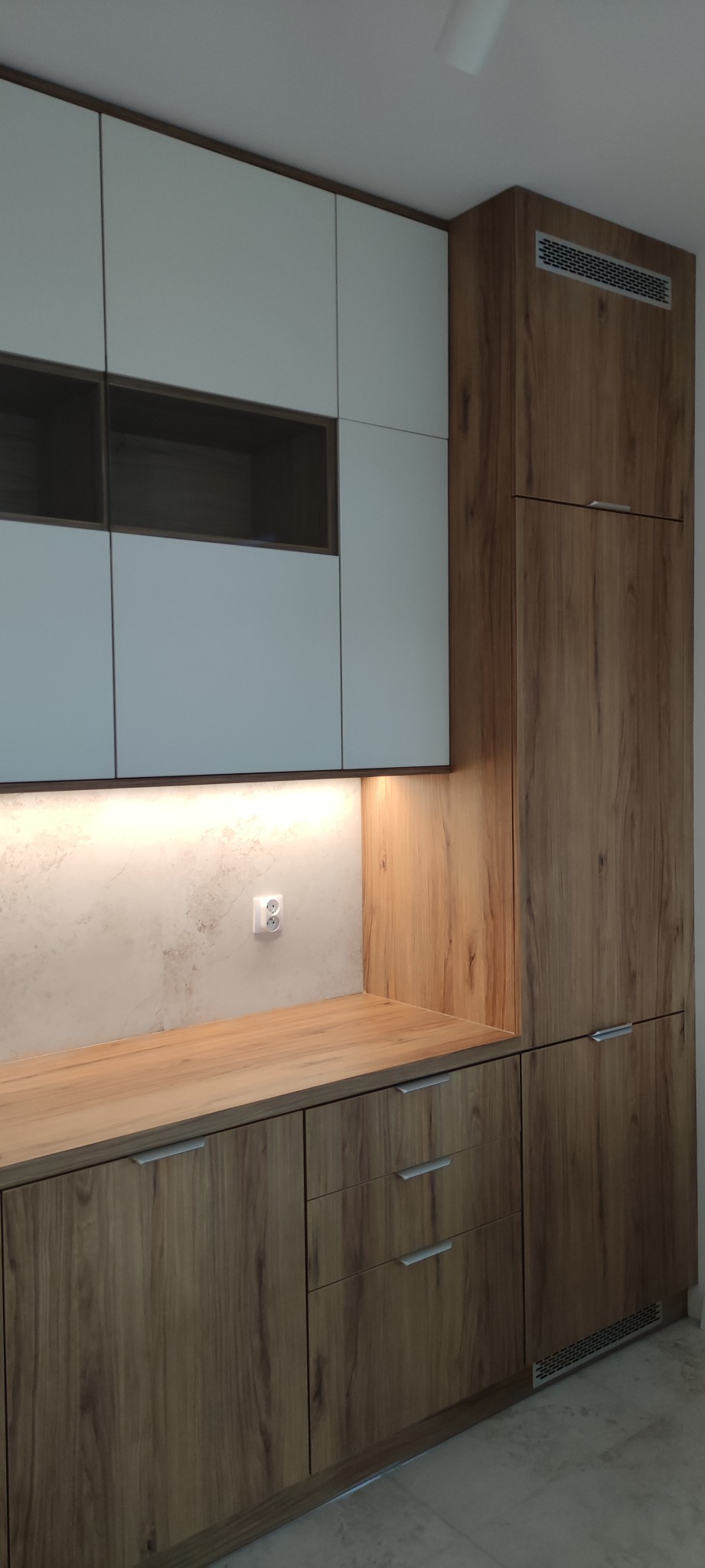 Nowoczesna zabudowa kuchenna z białymi szafkami górnymi i drewnopodobnymi szafkami dolnymi, oświetlenie LED podszafkowe, gniazdko elektryczne na ścianie.