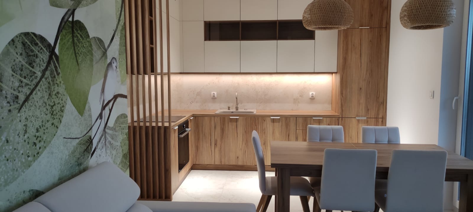 Nowoczesna kuchnia z drewnianymi szafkami i białymi górnymi szafkami, oddzielona od salonu drewnianą ażurową ścianką z motywem liści na ścianie, oświetlona lampami z wikliny nad drewnianym stołem...