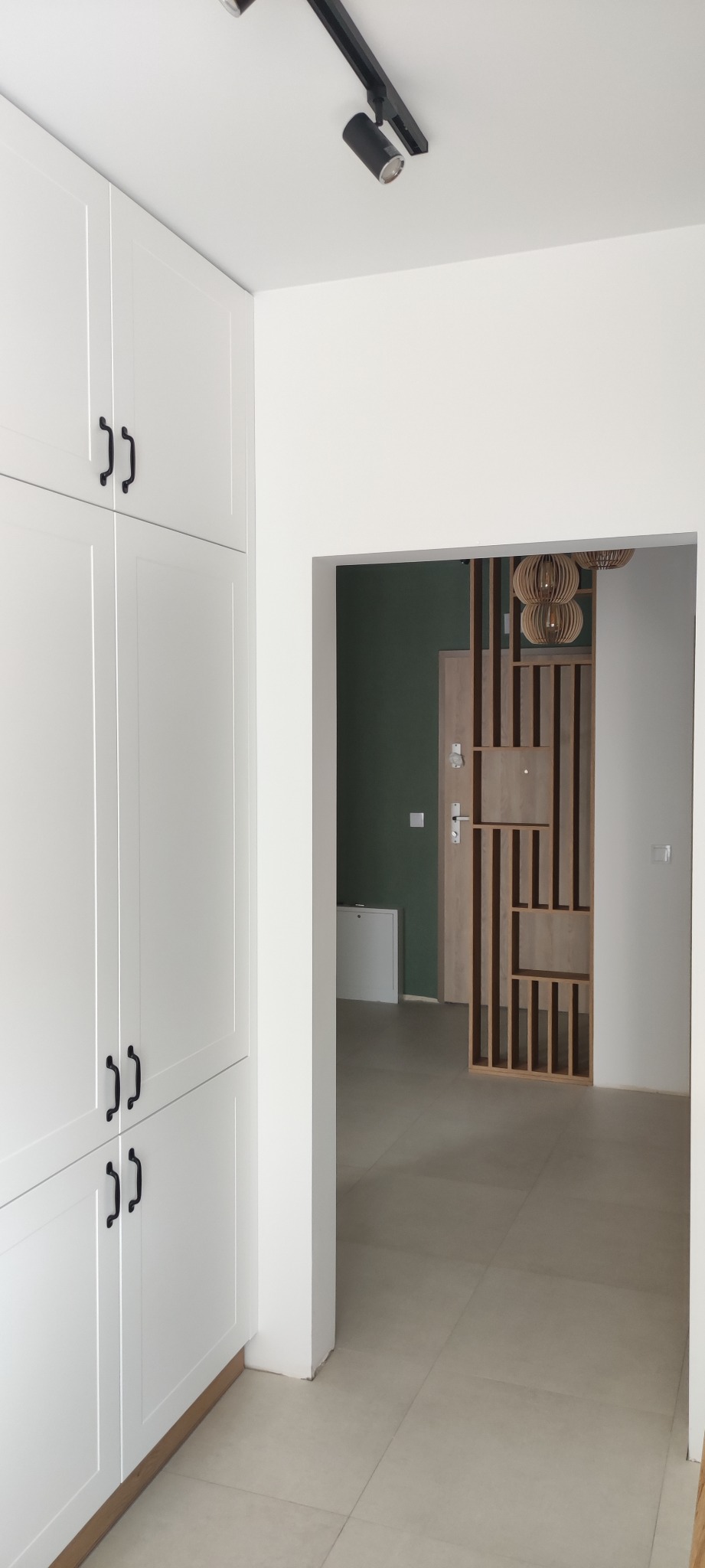 Wnętrze mieszkania z białymi szafami na ścianie, czarnymi uchwytami, widok na przedpokój z drewnianą ażurową ścianką działową i lampami w stylu boho.