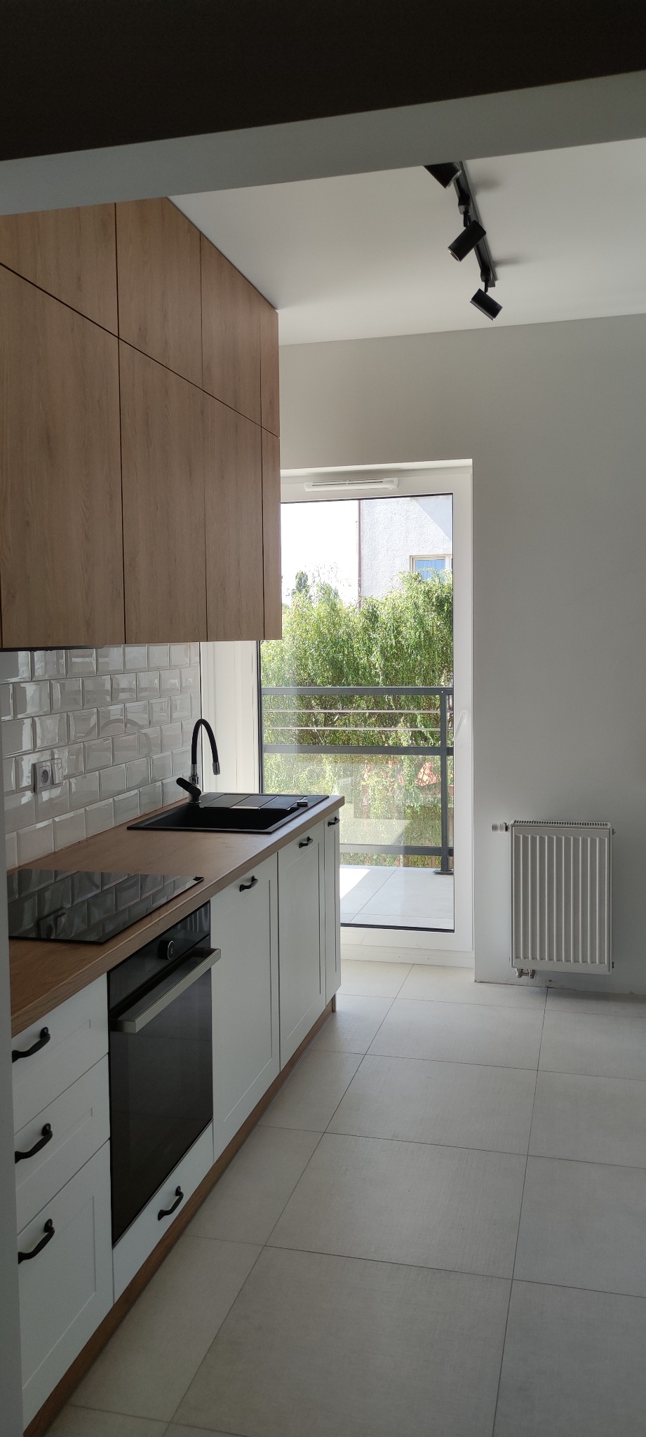 Jasna kuchnia z drewnianymi szafkami górnymi, białymi dolnymi, czarnym zlewem i płytą indukcyjną, widok na balkon i zieleń za oknem.