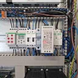 Usługi elektryczne F.W. - Szczegółowe ujęcie skrzynki elektrycznej z widocznymi bezpiecznikami, przekaźnikami czasowymi (ustawiony na 15:48:20), zasilaczem impulsowym Mean Well MDR-40-24 i licznymi przewodami w kolorach...
